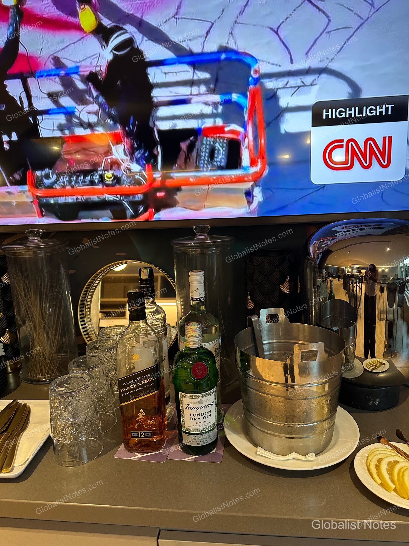 The liquor selection in the Hyatt Regency Malta lounge, featuring bottles of Johnnie Walker Black Label and Tanqueray, glassware, an ice bucket, and lemon slices, with a TV playing CNN in the background.