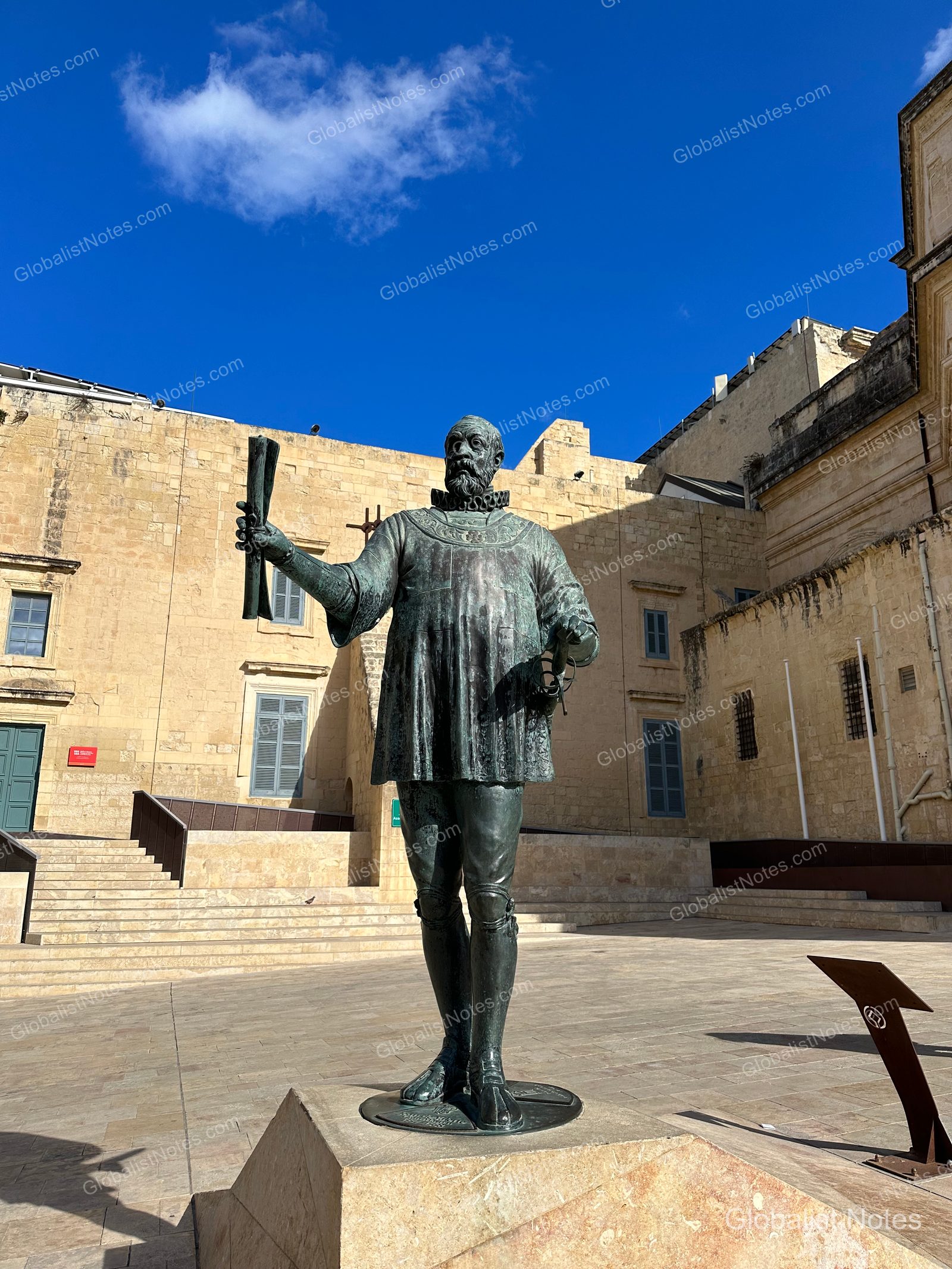 A bronze statue of a historical figure stands in a square under a clear Maltese sky, framed by antique stone buildings.