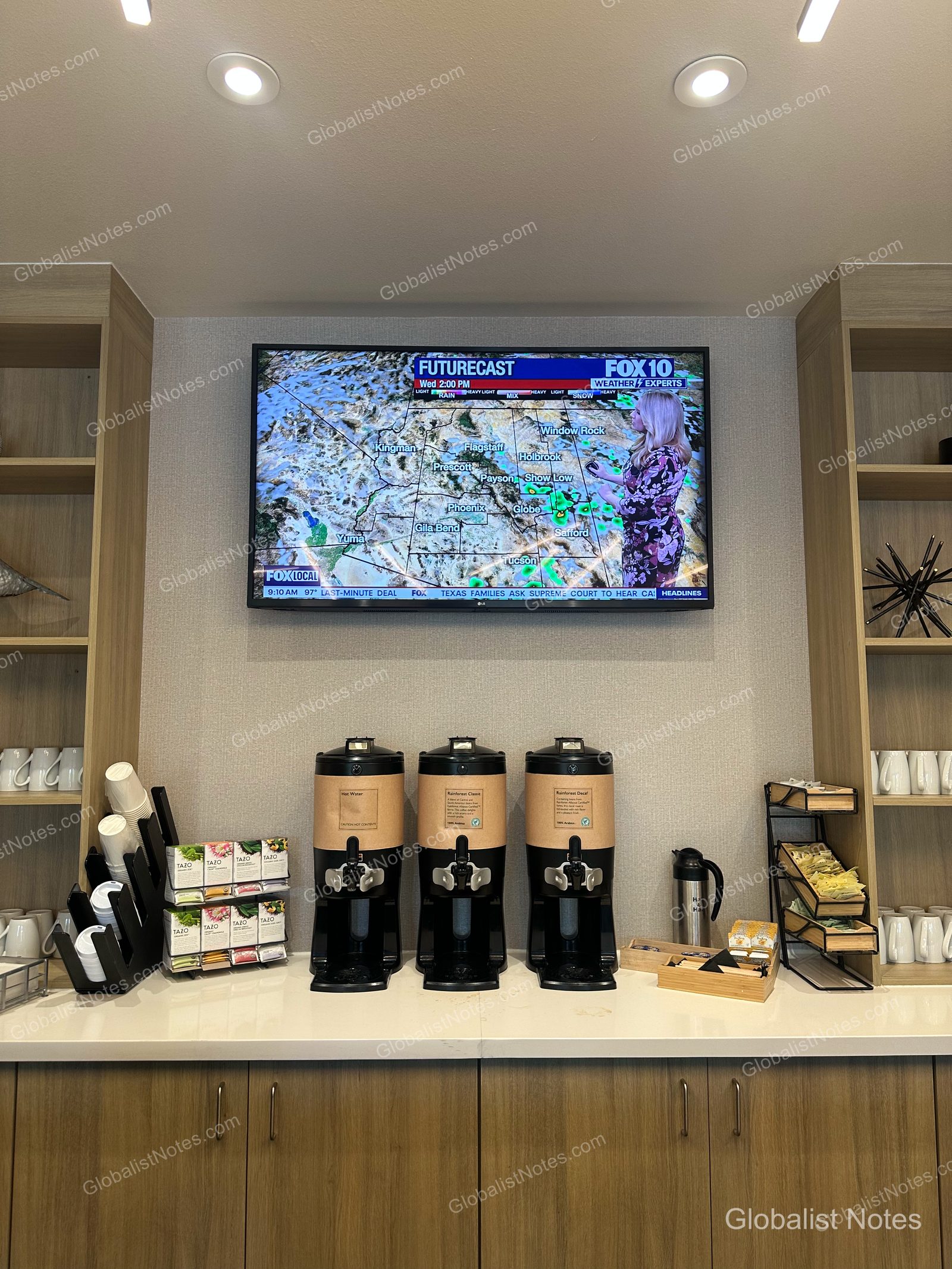 Hyatt Place Flagstaffの朝食エリアにあるコーヒーステーション。壁のモニターにはアリゾナ州の天気予報が表示され、その下に熱湯、コーヒーディスペンサー、Tazoのティーバッグ、紙コップなどが整えられています。