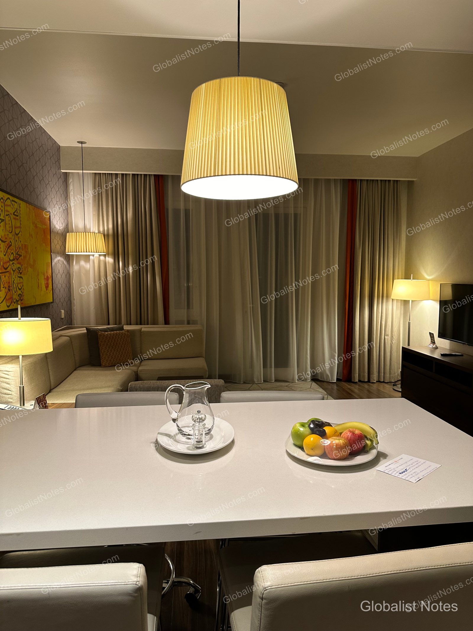 Hyatt Place Dubai Wasl District 一房套房的客廳。白色餐桌上擺放著迎賓水果與瓶裝水，後方可見米色沙發、燈具及落地窗簾。