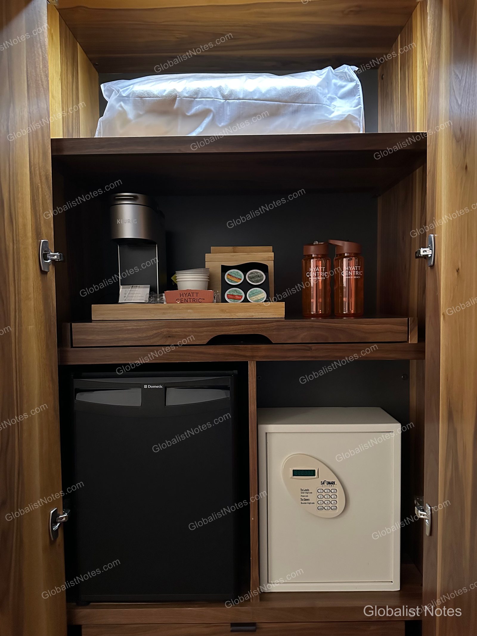 In-room storage unit at Hyatt Centric Waikiki Beach. The top shelf holds extra pillows, the middle shelf features a Keurig coffee maker with pods and two Hyatt Centric branded water bottles, and the bottom section houses a Dometic mini-fridge and an electronic safe side-by-side.