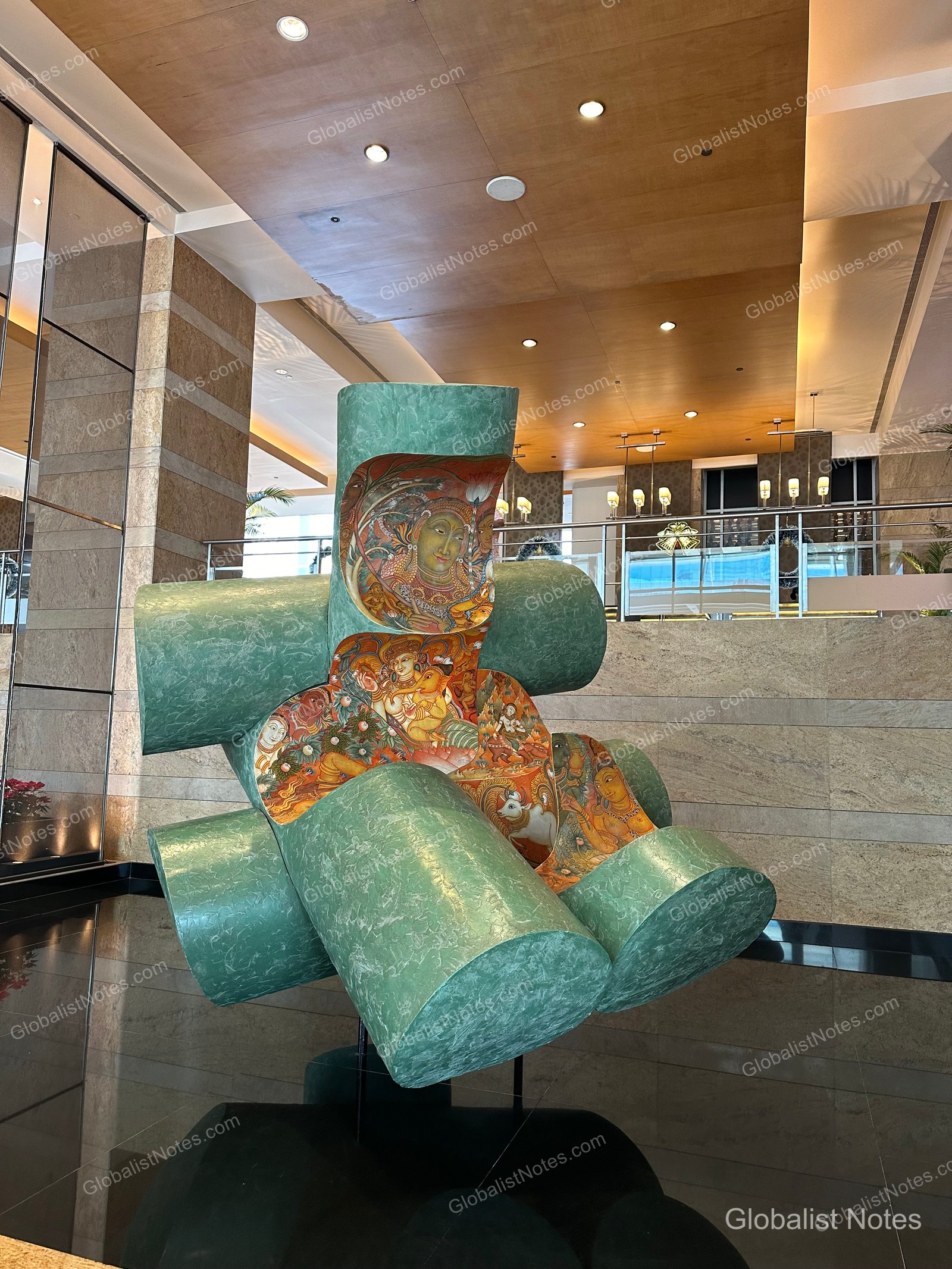 A large, mint-colored cylindrical installation in the center of the Grand Hyatt Mumbai lobby, featuring intricate traditional Indian paintings of figures and animals, set against the backdrop of the hotel's soaring ceilings.