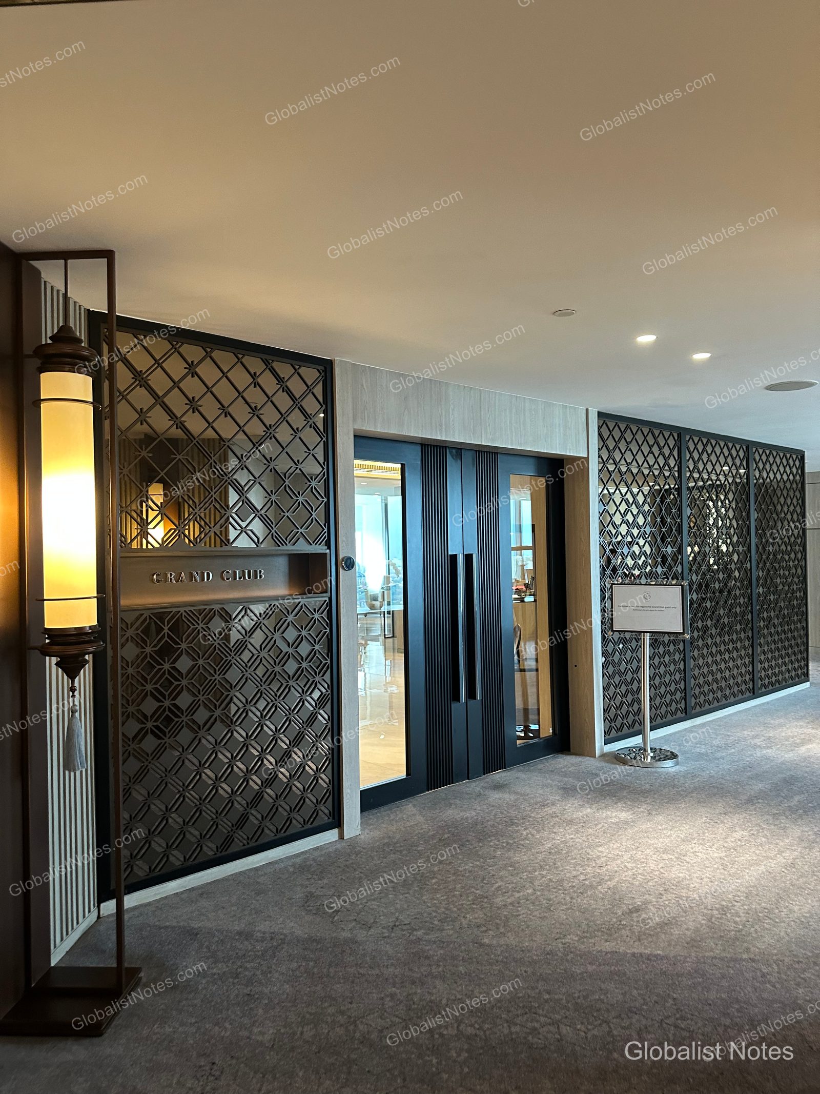 Entrance to the Grand Club Lounge at Grand Hyatt Jakarta. The glass doors are located at the end of a hallway featuring dark geometric metal latticework and sophisticated lighting, with an information sign standing near the entrance.