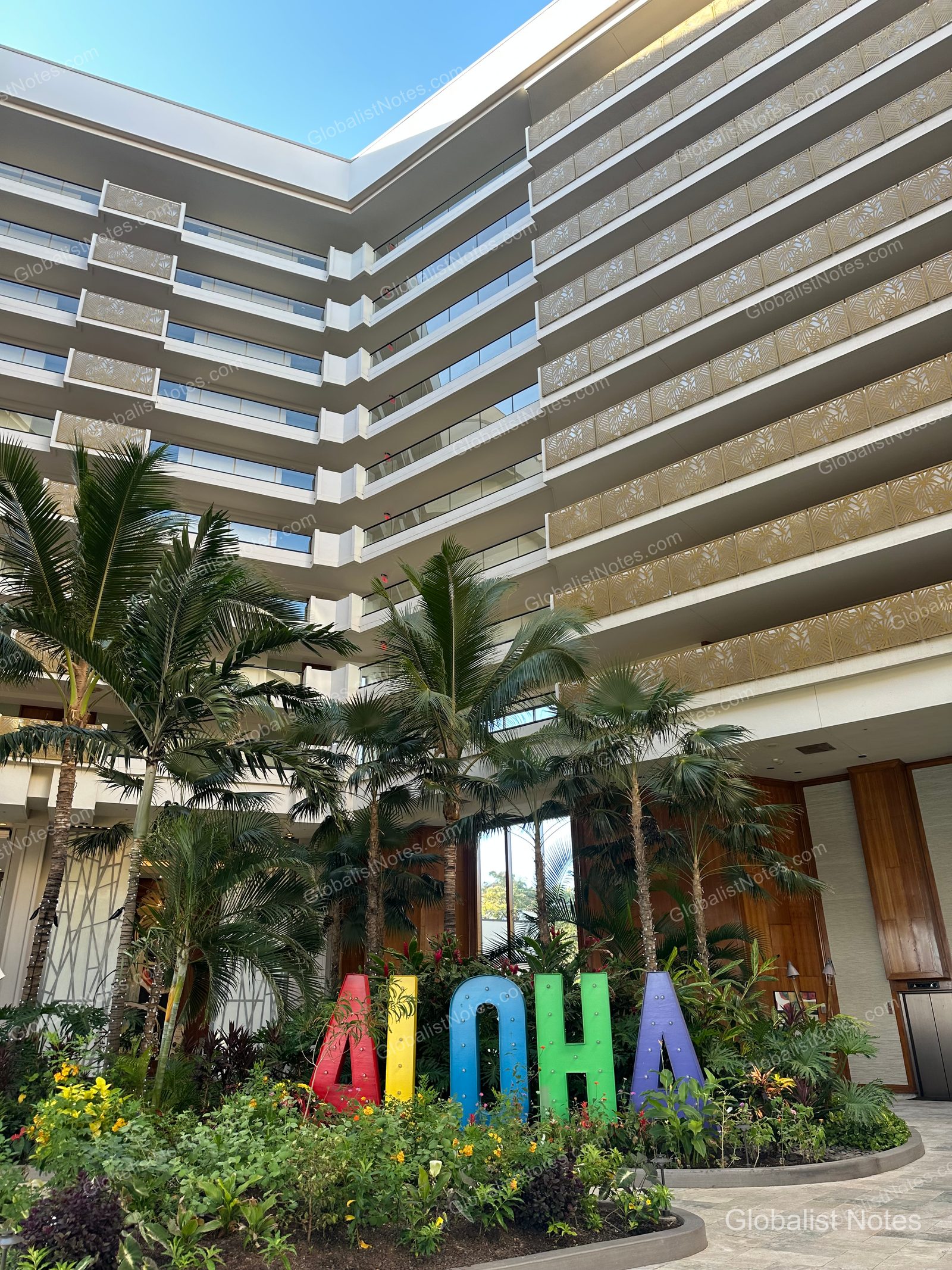 Exterior view of Hyatt Regency Maui Resort and Spa in Lahaina, Hawaii, featuring a multi-story building with balconies, a garden in front with a colorful 'ALOHA' sign, surrounded by palm trees and flowers.