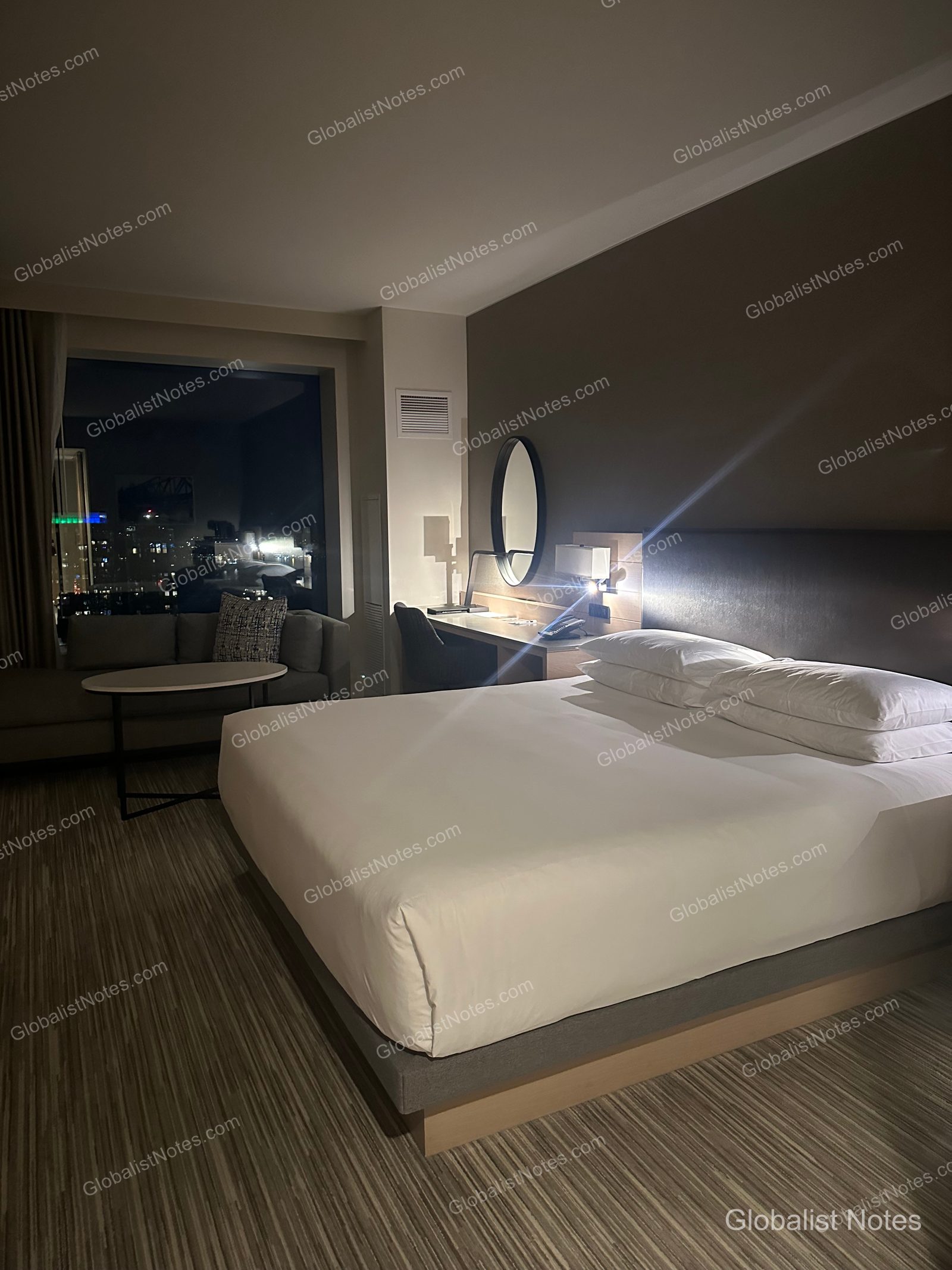 Interior of a standard king room at Hyatt Regency Seattle. A king bed with white linens is centered in the room, with a sofa and table placed by the window. The desk lamp is on, and the Seattle city skyline is visible at night through the window.