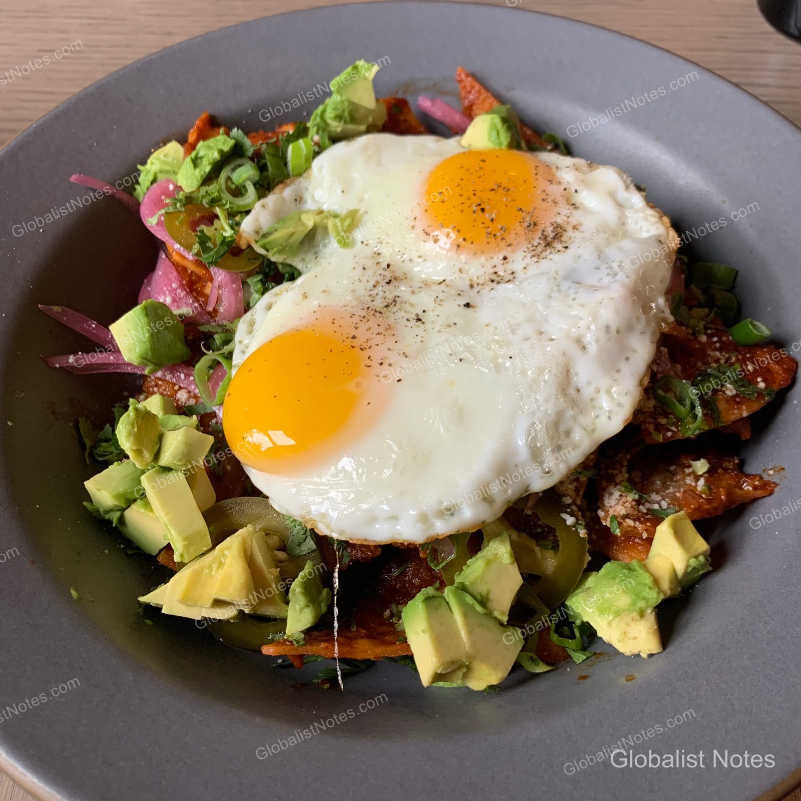 Chilaquiles served on a gray plate, featuring two fried eggs over crispy tortilla chips, garnished with diced avocado, pickled onions, and scallions.