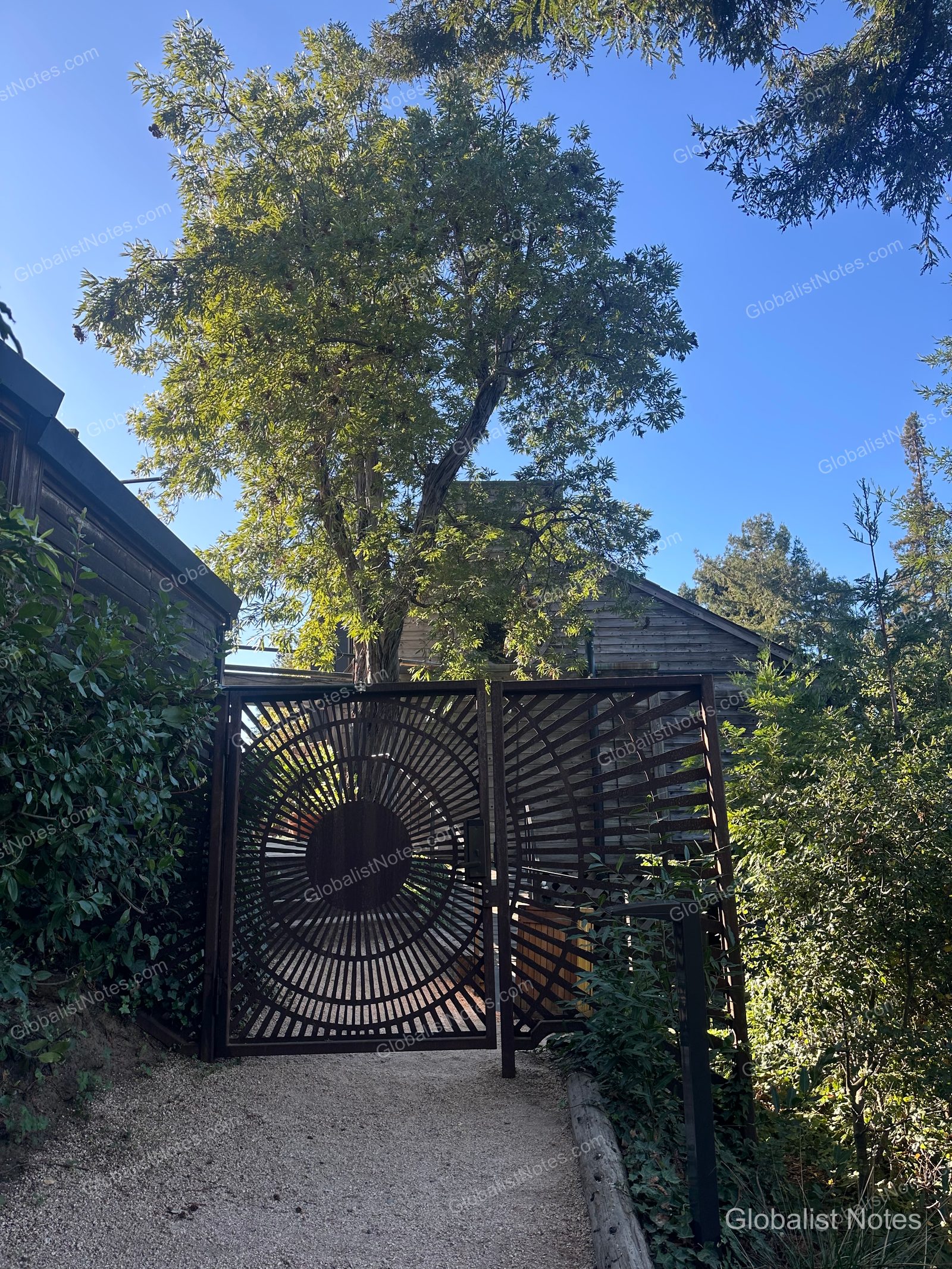 The entrance to Alila Ventana Big Sur, featuring a unique sun-shaped metal gate at the end of a narrow path, with dense trees and wooden buildings set against a blue sky.