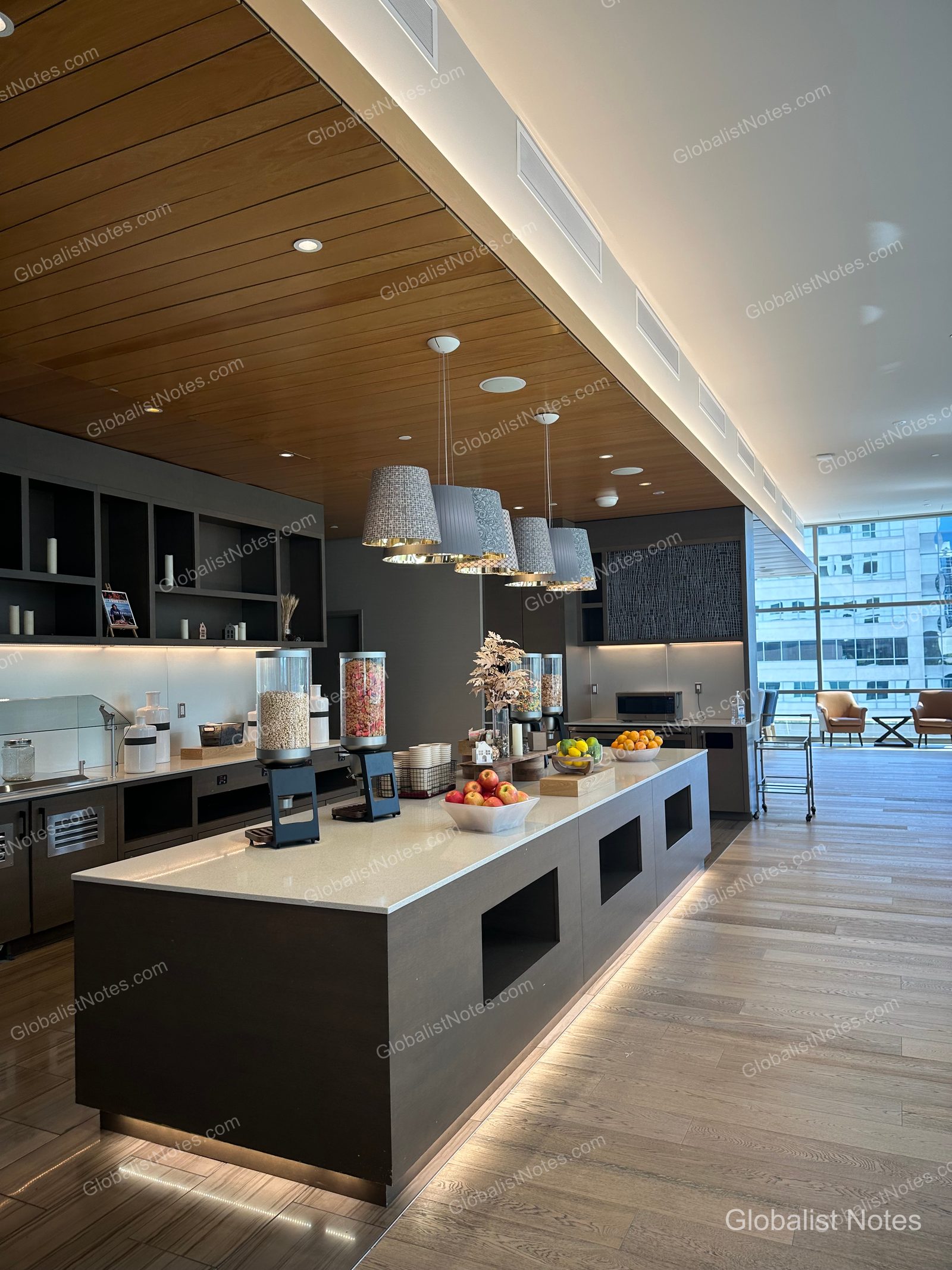 Interior view of the Regency Club lounge at Hyatt Regency Seattle. A long central island features cereal dispensers and a fruit basket, with wood paneling and pendant lighting on the ceiling. Seattle city buildings are visible through the windows, and the floor is finished in light-toned wood.