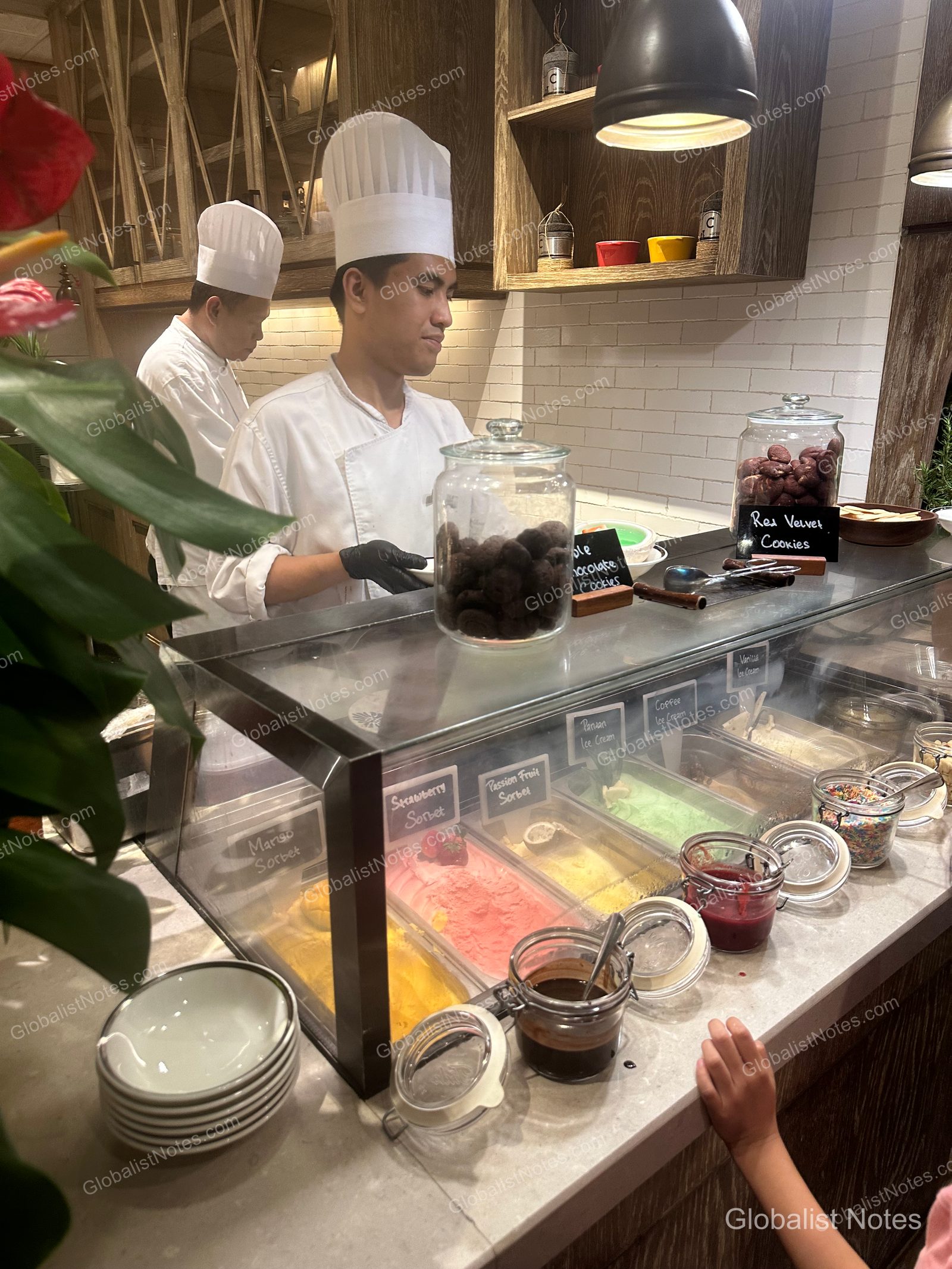 Two chefs in white uniforms stand behind the ice cream station at the Grand Hyatt Jakarta breakfast buffet. The glass display case contains mango, strawberry, and passion fruit sorbets, along with various cookies.
