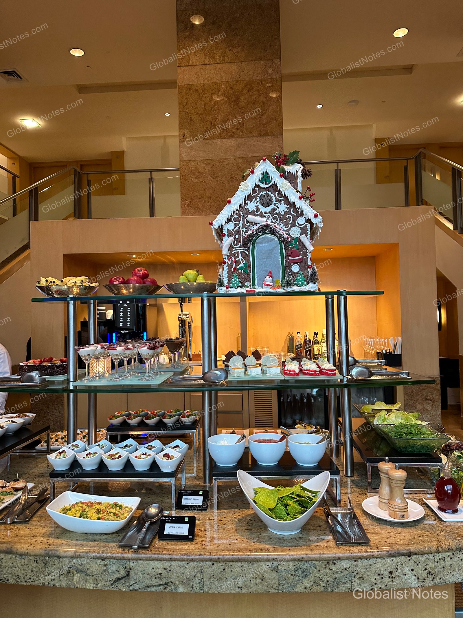 A buffet station in the Grand Hyatt Mumbai lobby. A gingerbread house sits on a glass shelf above an assortment of salads, desserts, and fruits, with bowls of nachos and sauces arranged below.