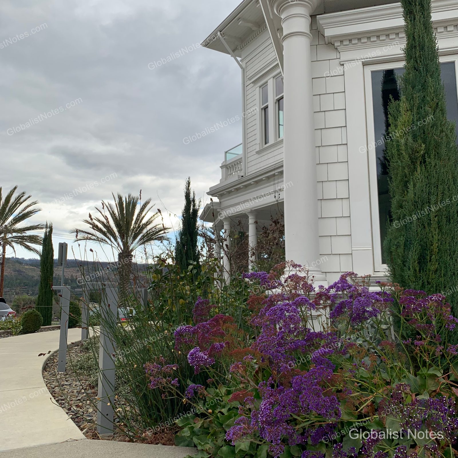 A white building with columns at Alila Napa Valley in St. Helena, adjacent to a garden in full purple bloom and an outdoor walkway under a cloudy sky.