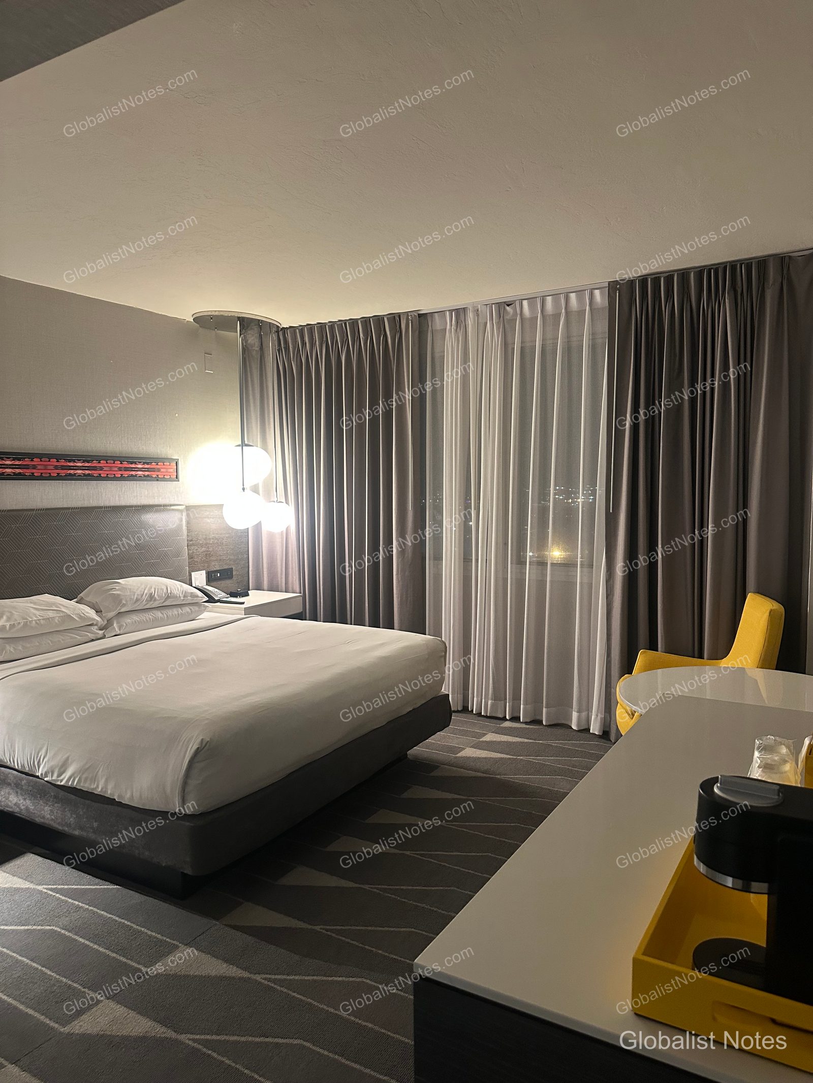 Interior of a One Bedroom King Suite at Hyatt Regency Los Angeles International Airport. On the left, a king bed with white linens and pendant lighting; on the right, a desk with a yellow chair, table, and coffee machine, with curtains drawn over the window.