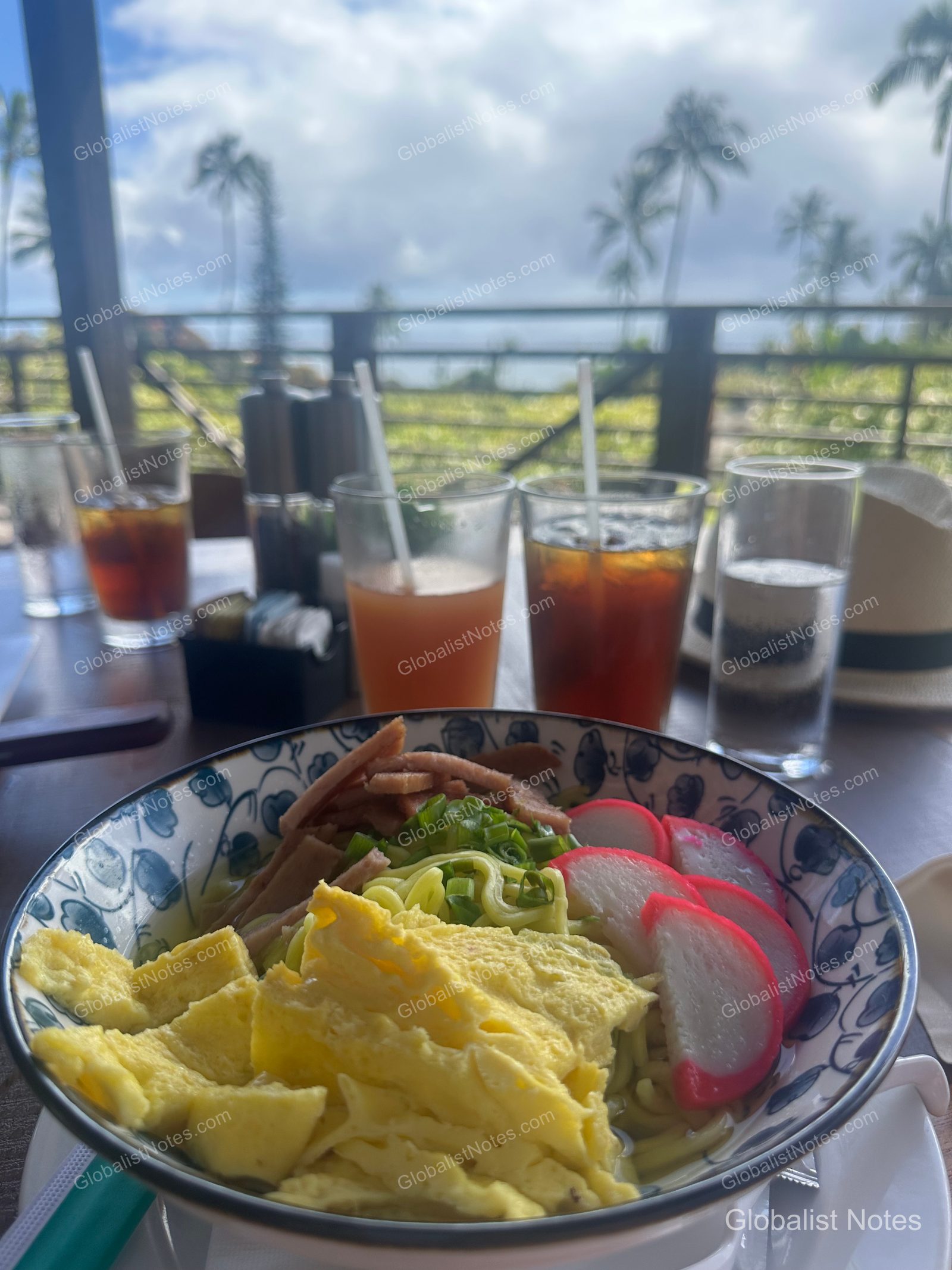 A bowl of ramen topped with egg garnish, fish cake, and meat, served alongside several drinks on an outdoor terrace table at Hana-Maui Resort, with a scenic view of palm trees and a cloudy sky in the background.