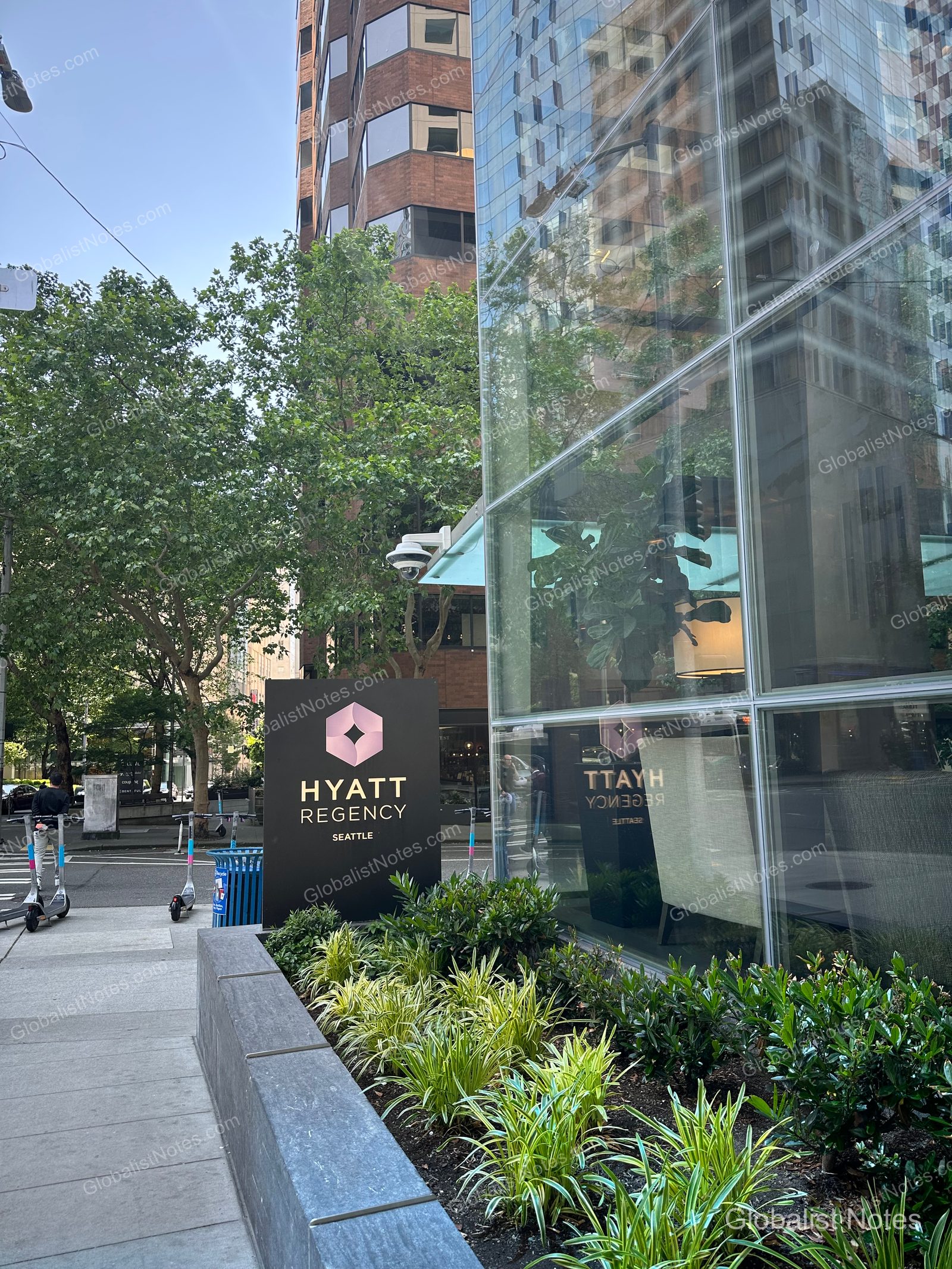 Exterior view of Hyatt Regency Seattle in the city center. A black hotel logo sign stands next to a flower bed, while the glass facade on the right reveals the lobby's greenery and lighting. Electric scooters are parked on the sidewalk.