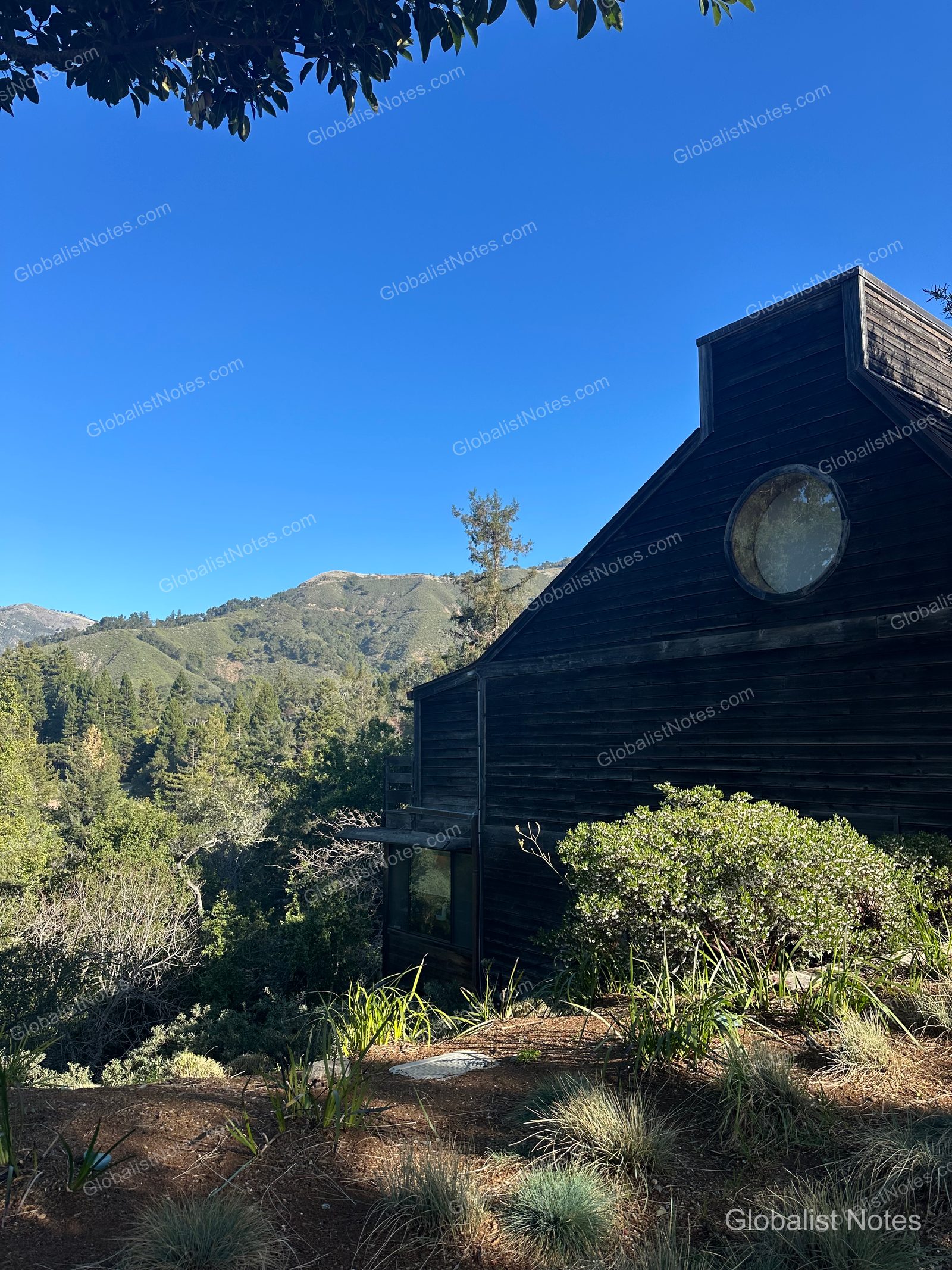 The dark wood exterior and circular windows of Alila Ventana Big Sur under a clear blue sky, surrounded by dense forest and mountain ranges.
