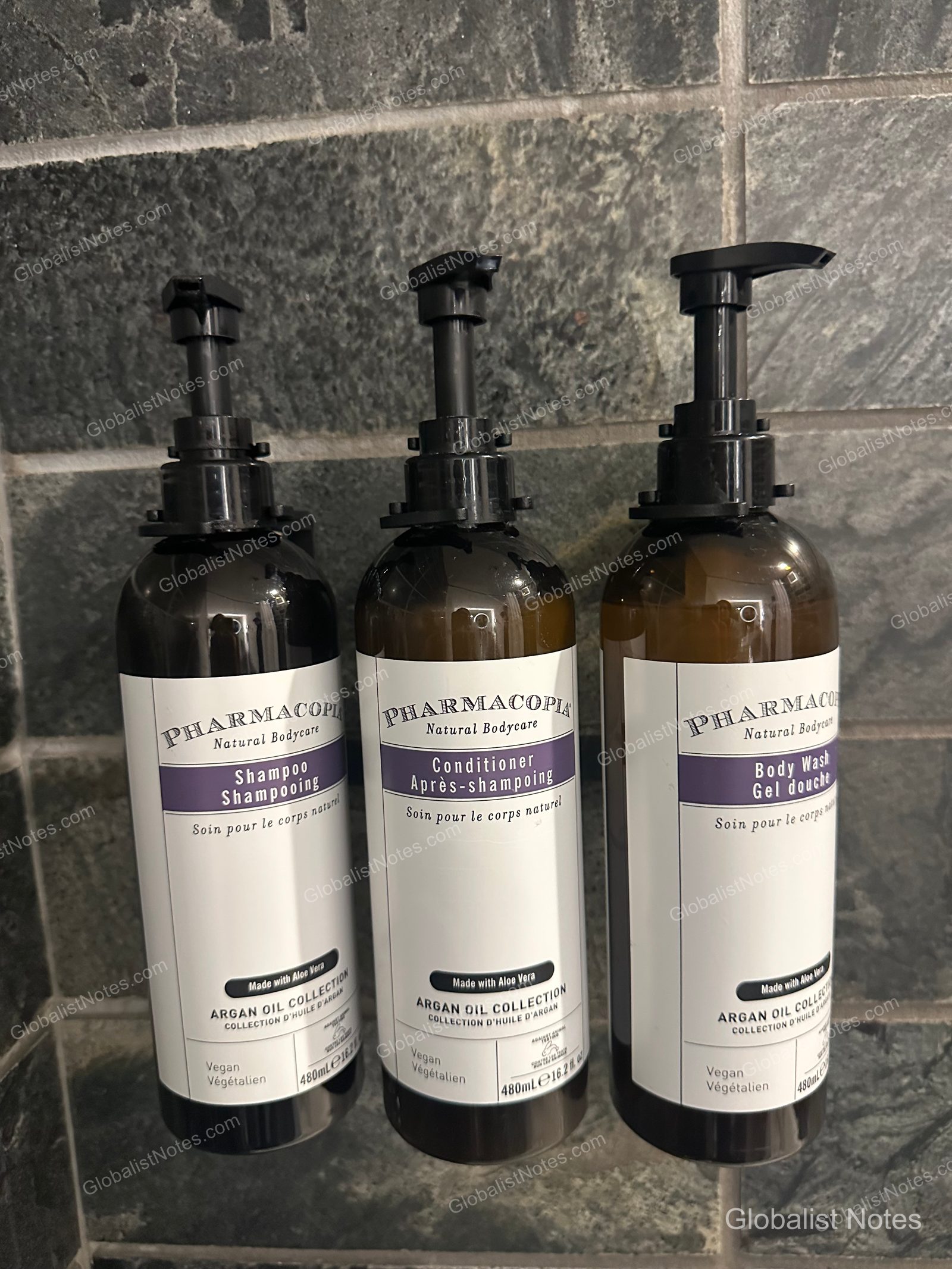 Pharmacopia Argan Oil collection amenities mounted on the bathroom wall of a Deluxe Suite at Hyatt Regency Vancouver. The pump-dispenser bottles for shampoo, conditioner, and body wash are lined up against a dark gray tiled wall.
