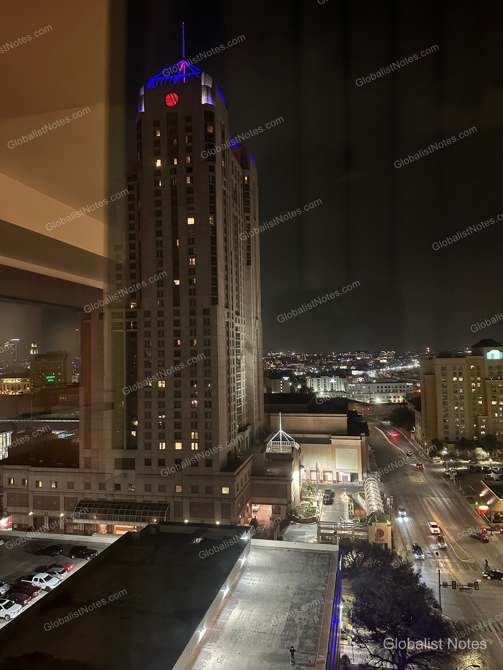 A nighttime view of the San Antonio River Walk, looking out from a window at the Grand Hyatt. The city skyline and the hotel's high-rise exterior are illuminated, with streets and parking areas visible below.