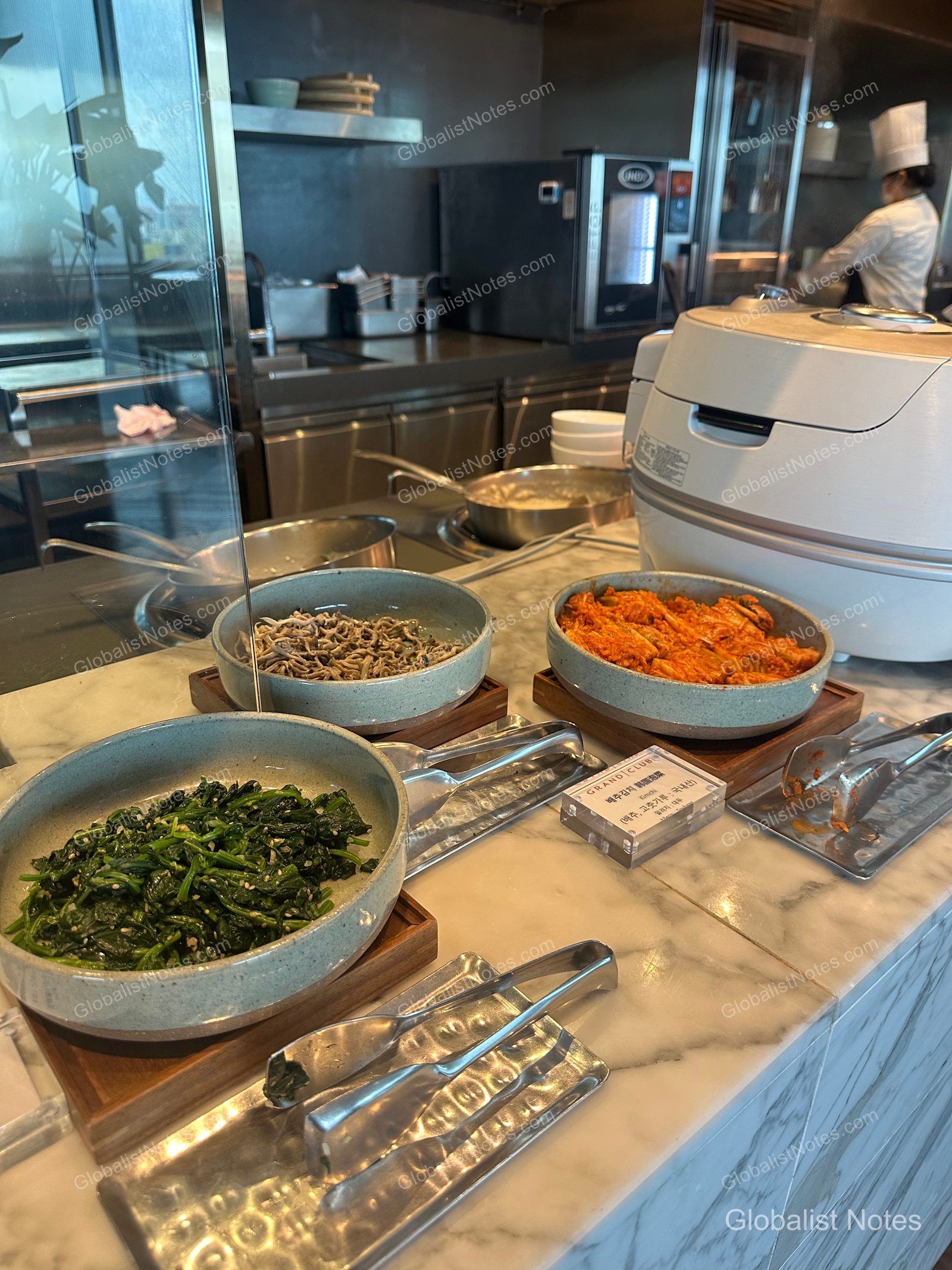 Korean side dishes including spinach, stir-fried anchovies, and kimchi displayed on the marble counter at the Grand Hyatt Jeju breakfast buffet, with chefs preparing food in the open kitchen in the background.