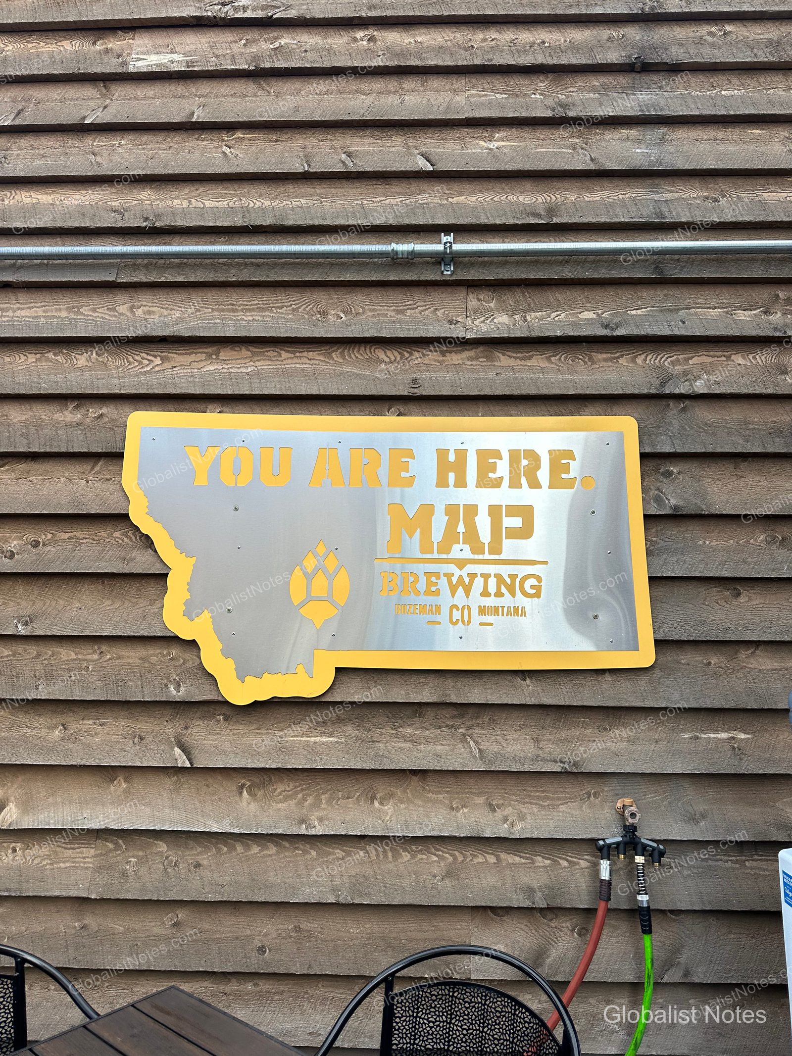 木壁に掲げられたモンタナ州の形をした金属看板。「YOU ARE HERE. MAP BREWING BOZEMAN CO MONTANA」の文字とホップのロゴが刻まれています。