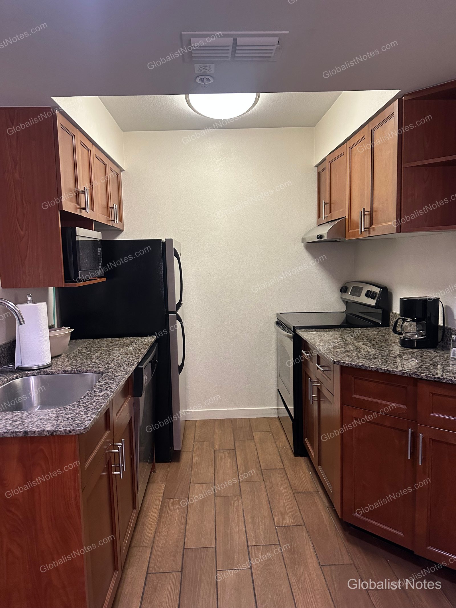 Hyatt House Scottsdale/Old Townの1ベッドルームスイートのキッチン。ブラウンの木製キャビネットに花崗岩のカウンタートップ、黒の冷蔵庫、オーブン、電子レンジ、コーヒーメーカーを備え、床は木目調のタイル張りです。