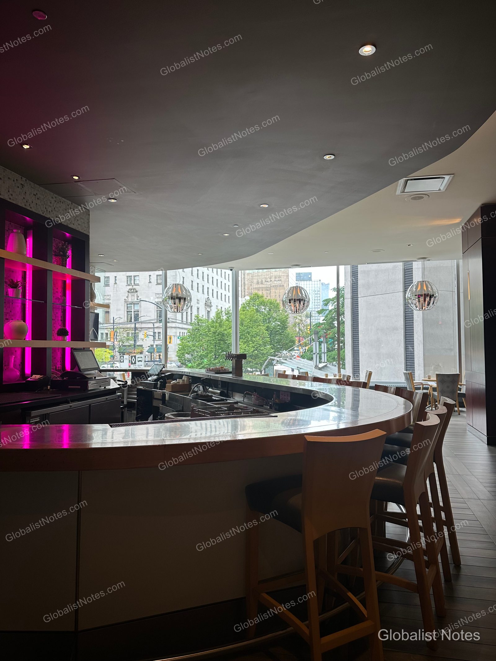Curved bar counter at Hyatt Regency Vancouver. The shelves behind the bar are illuminated with pink LED lighting, and the floor-to-ceiling windows offer a view of the downtown Vancouver cityscape and street trees. High stools are arranged in front of the bar, with recessed ceiling lights and pendant fixtures overhead.