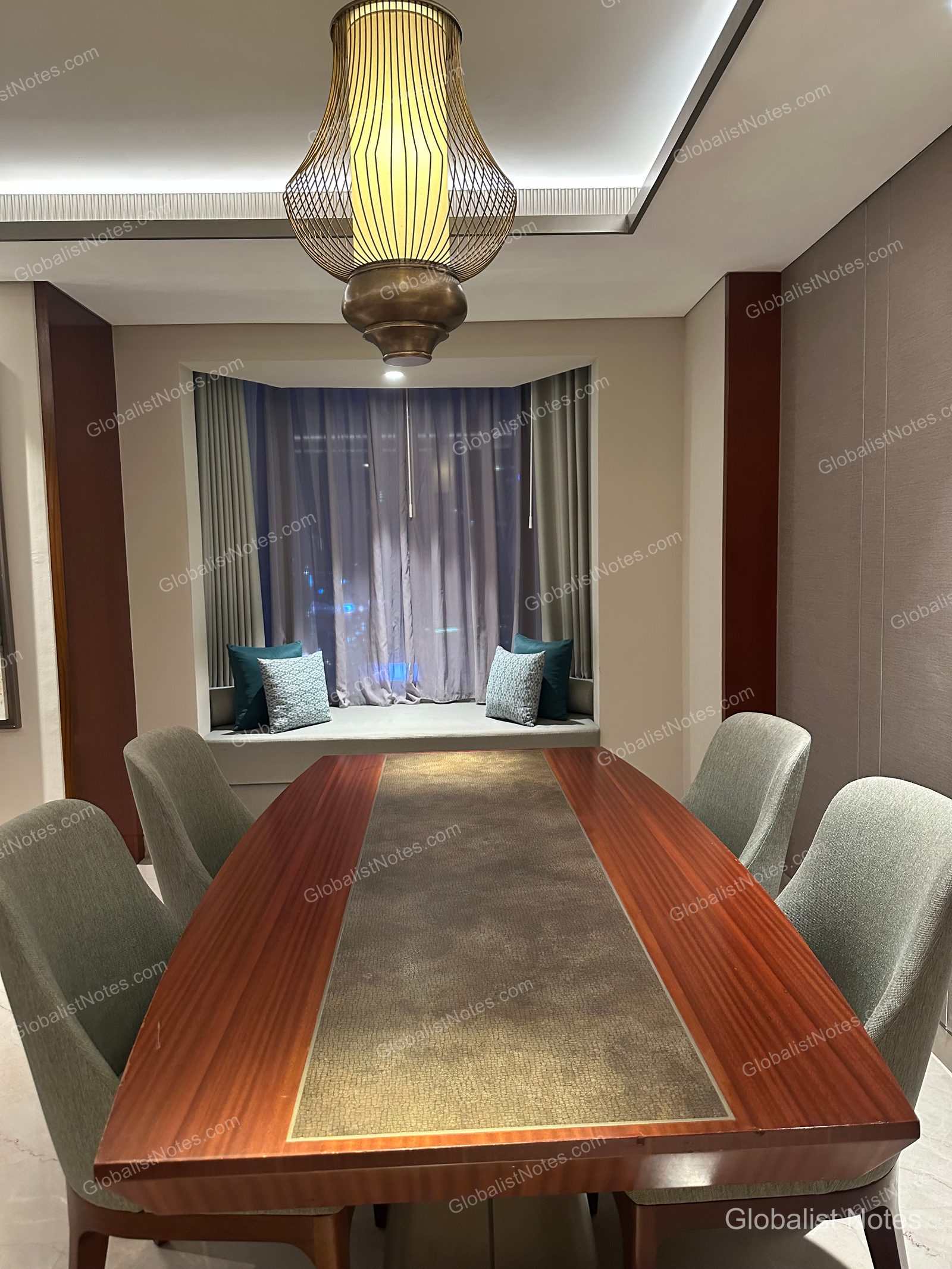 Dining area of a Grand Suite at Grand Hyatt Jakarta. A long wooden dining table with four gray chairs is centered under a unique metal frame light fixture. A cushioned window seat is set against the window, with the faint Jakarta night skyline visible through the curtains.