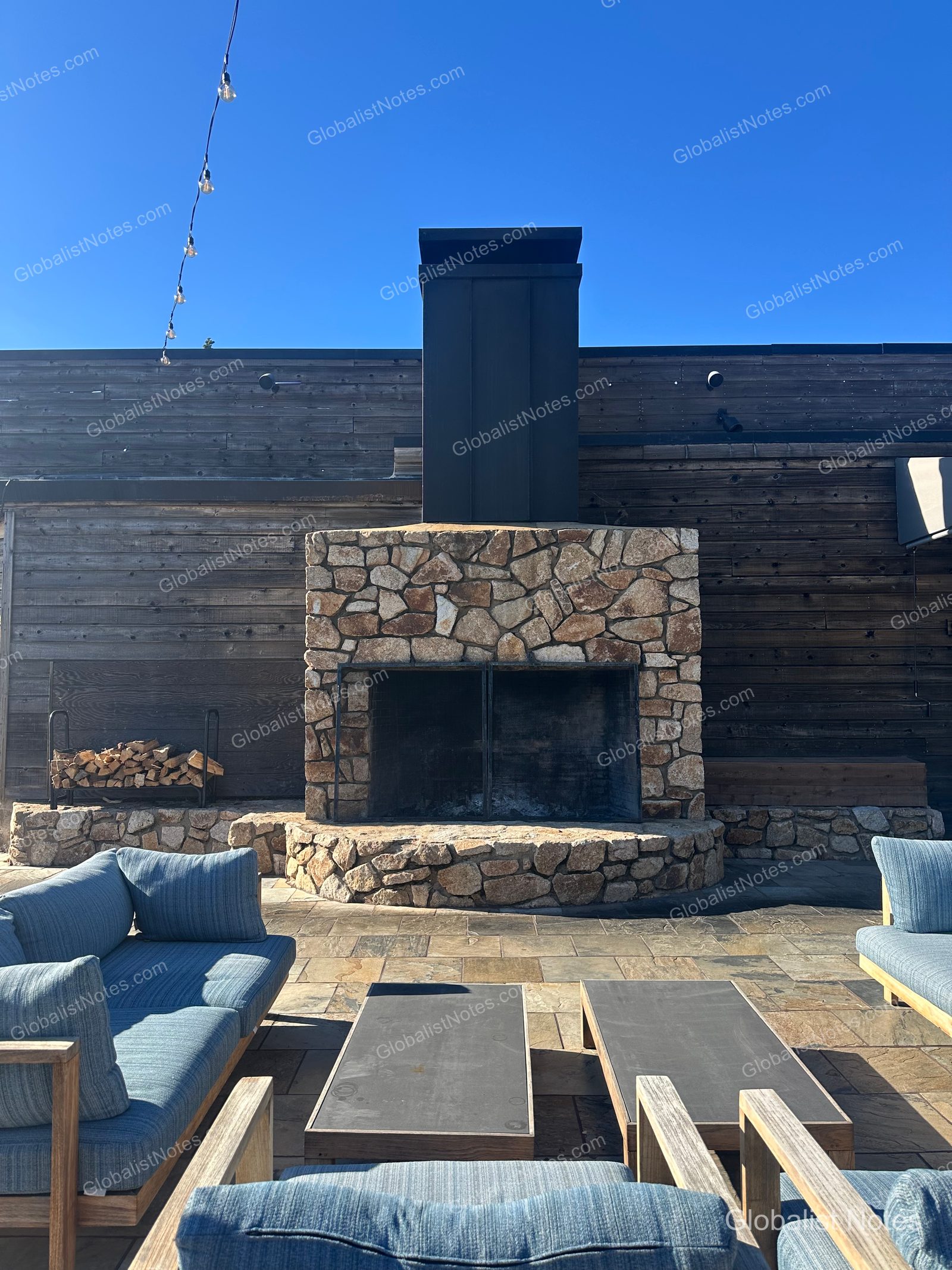 A lounge area on the outdoor terrace at Alila Ventana Big Sur with a large stone fireplace, blue sofas, and a wooden table. The wall is finished with dark wood, string lights hang above, and firewood is stacked to the side.