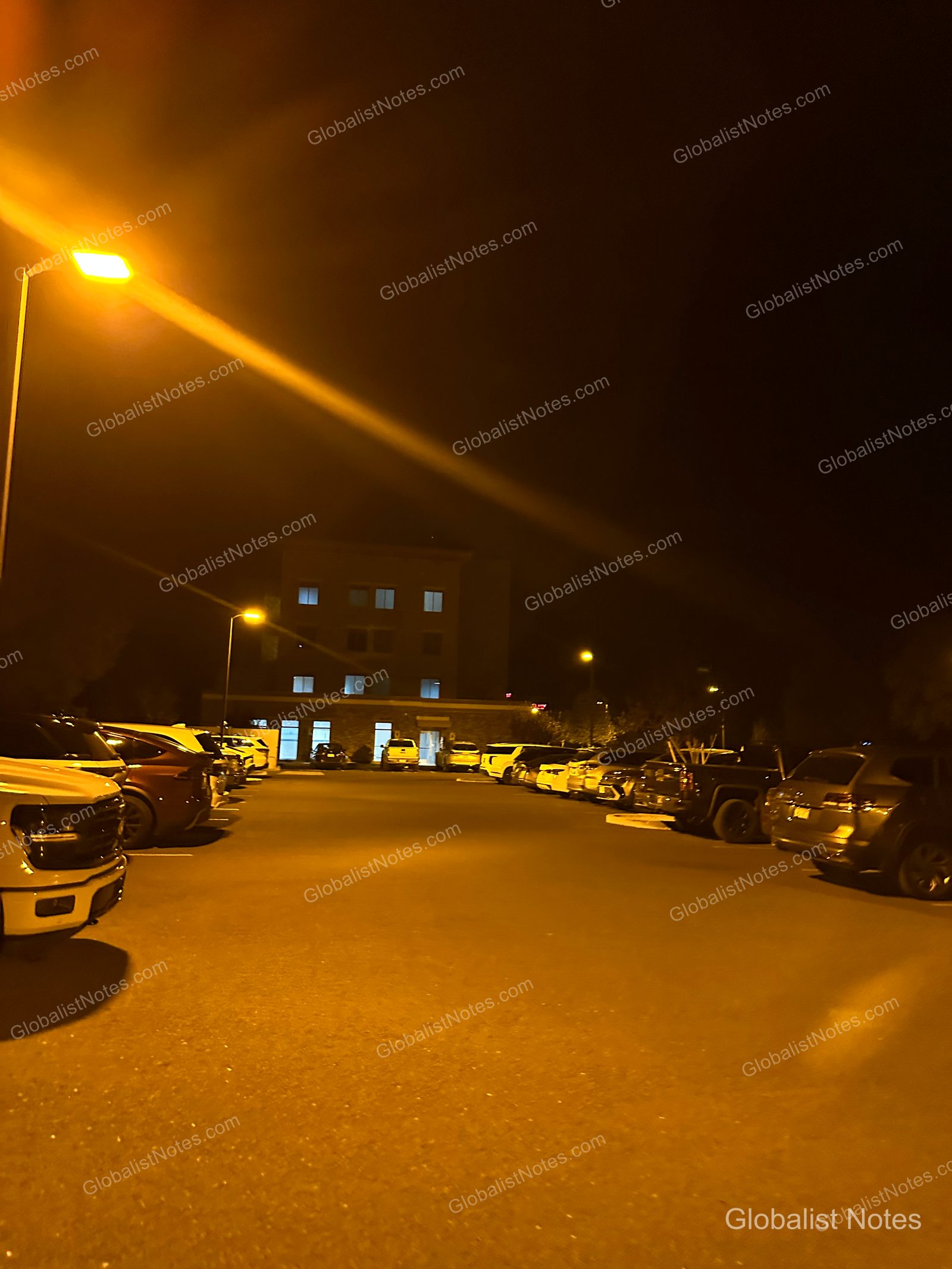 夜空の下、街灯に照らされたHyatt Place Flagstaffの屋外駐車場。駐車車両が並び、遠くにホテルの明かりが灯った窓が見える。
