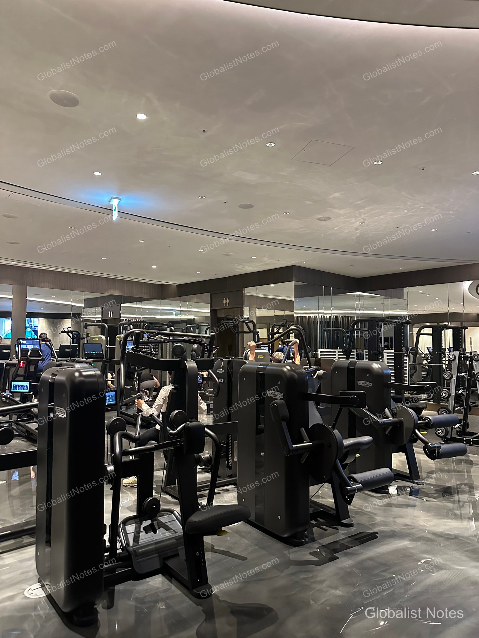 Interior of the fitness center at Grand Hyatt Jeju, featuring black Technogym strength training equipment spaced evenly throughout the room, with guests working out. The ceiling has recessed lighting, and the floor is finished in a polished, dark-toned material.