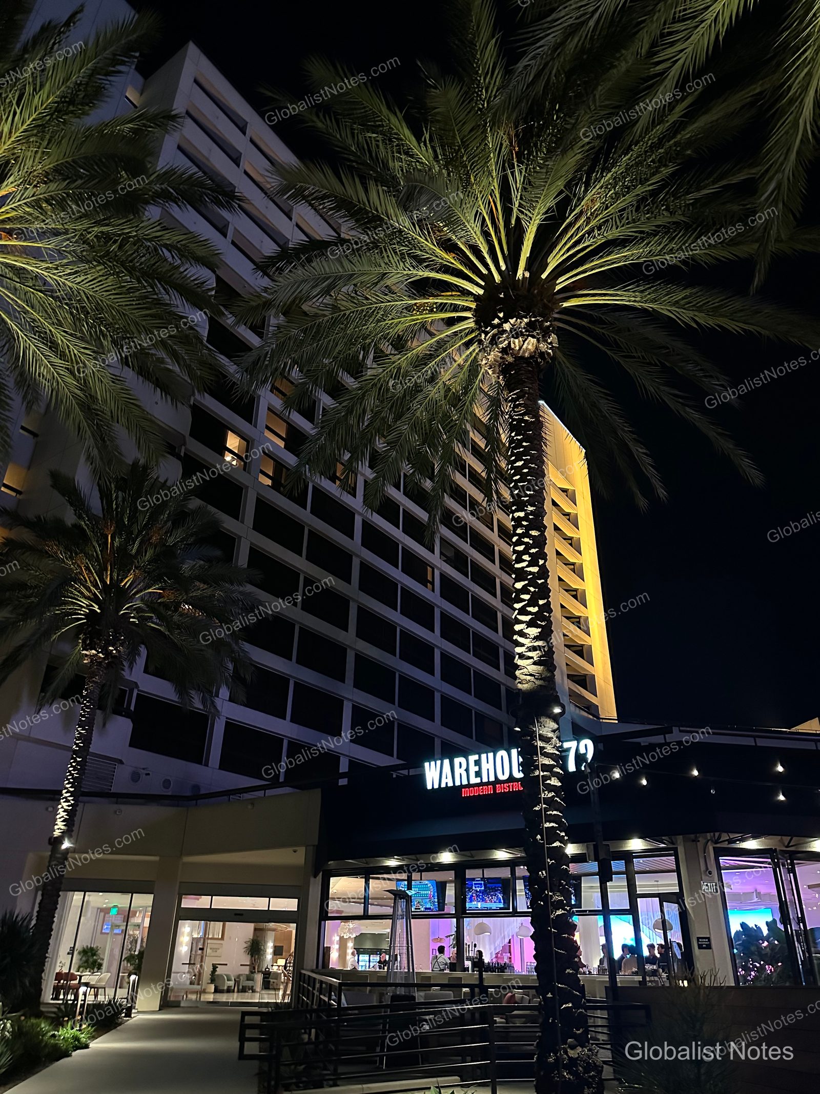 夜空下點燈的 Hyatt Regency Irvine 外觀，以及位於入口處的 Warehouse 72 餐廳。建築前方矗立著高大棕櫚樹，餐廳明亮的燈光透過玻璃窗映照出來。