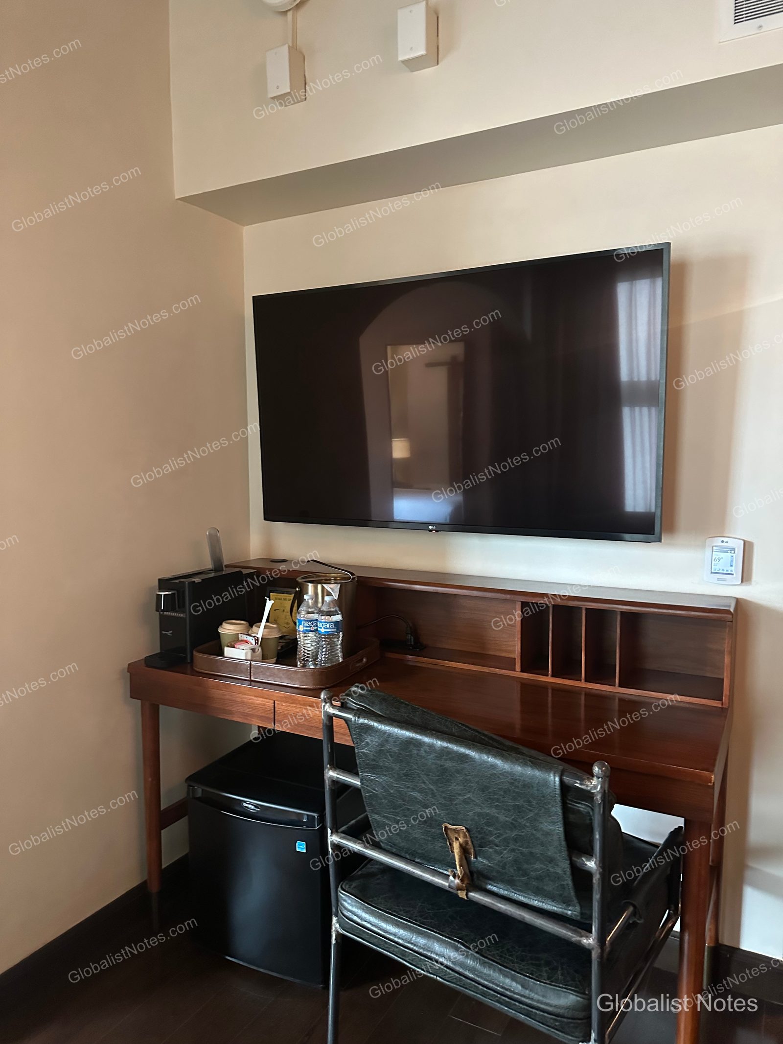 Inside the Junior Suite at Hotel Figueroa in Los Angeles, featuring a wall-mounted TV above a wooden desk equipped with a coffee machine, bottled water, and a mini-fridge, accompanied by a vintage leather chair.