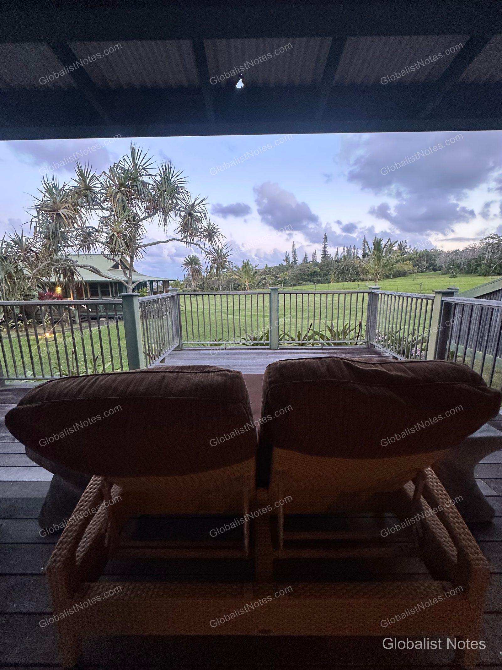 A peaceful view of Maui's green meadows, tropical trees, and the distant ocean, seen past two rattan chairs on the terrace of a 1 King Bed Ocean View Bungalow at Hana-Maui Resort.