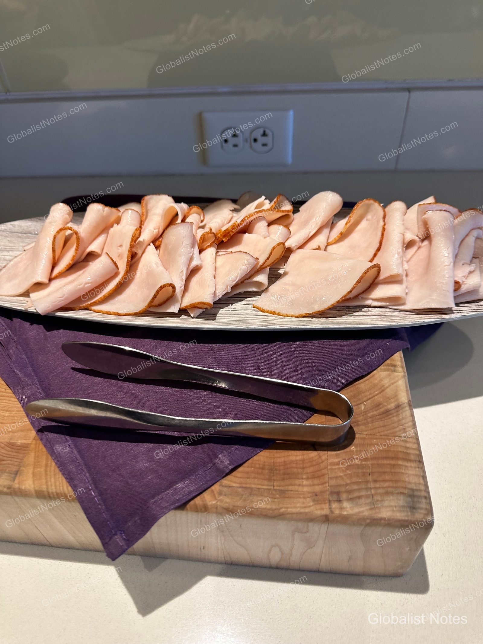 A plate of thinly sliced turkey ham at the Hyatt Regency Maui Resort and Spa breakfast buffet, set on a wooden cutting board with a purple napkin underneath and metal tongs provided.