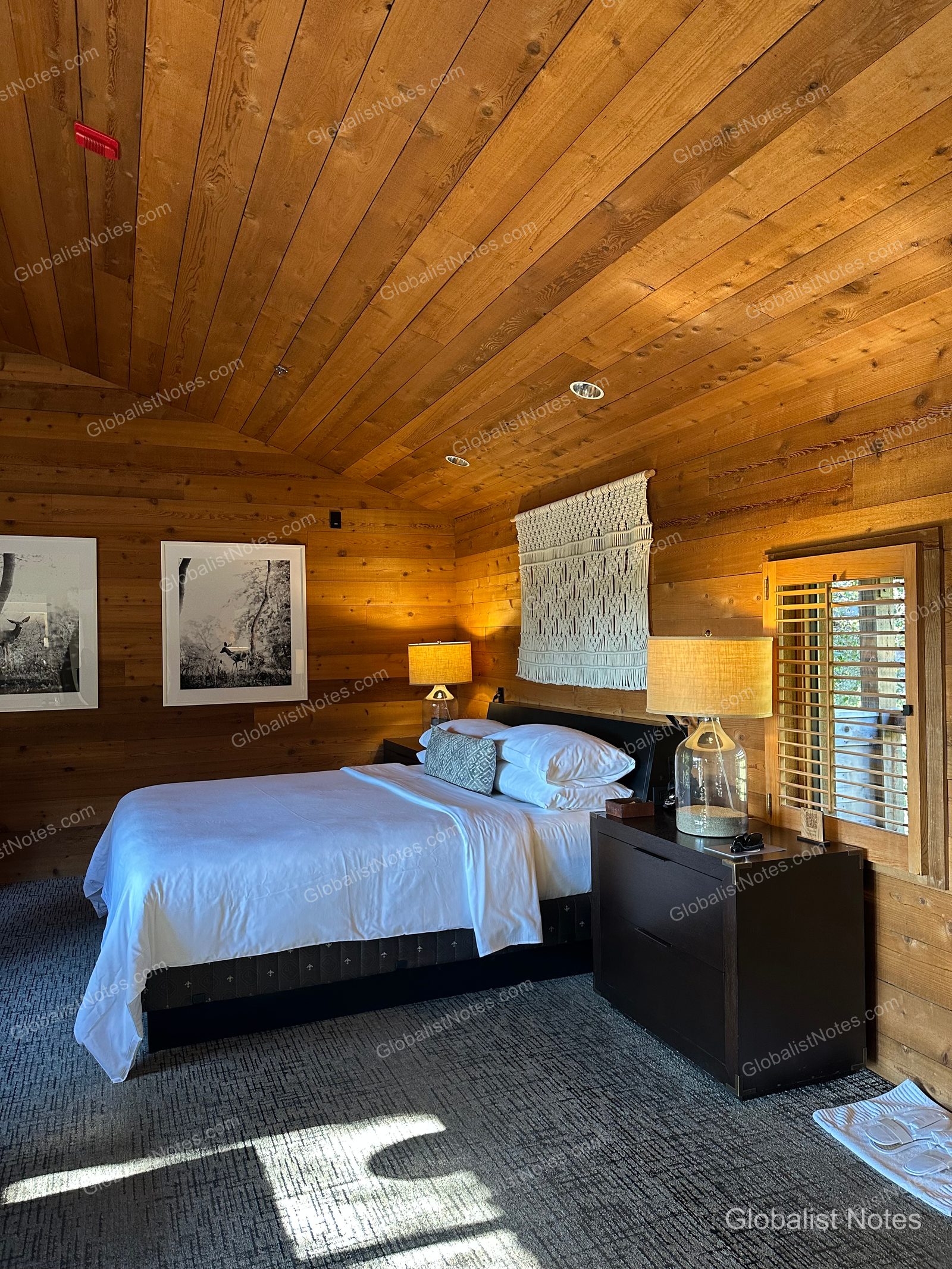 Interior of a Junior Suite at Alila Ventana Big Sur. The room features wood walls and ceilings, a queen bed with side tables and lamps, macramé wall art, black-and-white framed photos, and a window with wooden shutters.