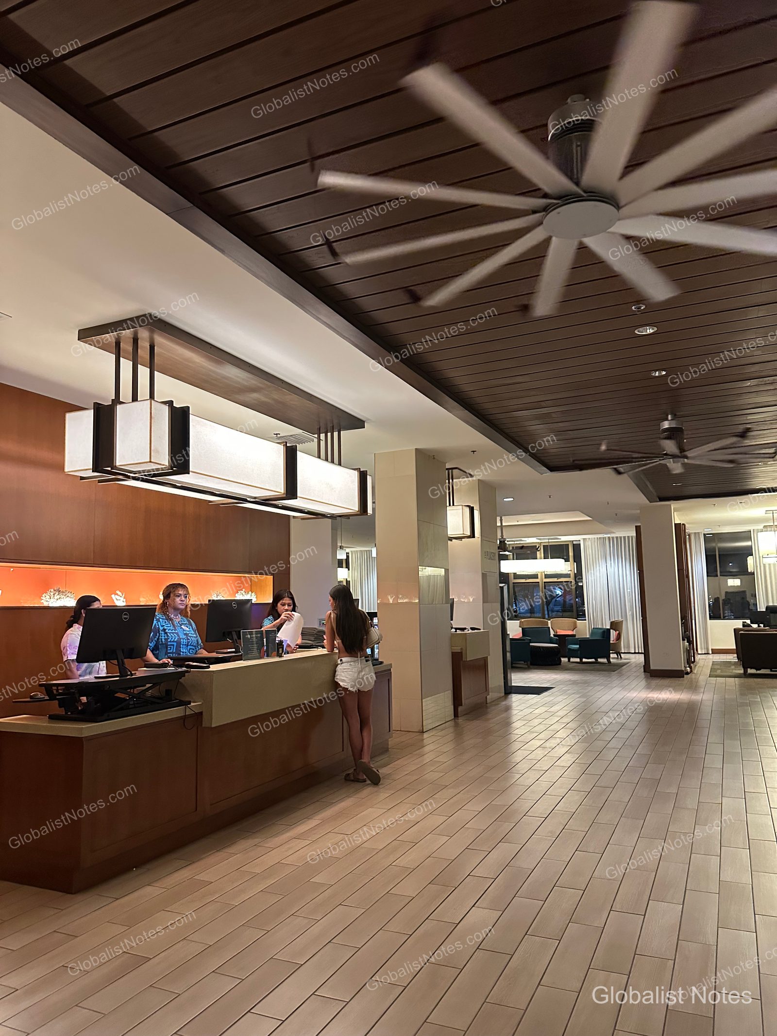 Lobby view of the Hyatt Place Waikiki Beach in Honolulu, Hawaii. A large ceiling fan rotates under a wood-paneled ceiling, with a long check-in counter and guests standing in front. The floor is finished with light-toned tiles, and a seating area is visible in the background.
