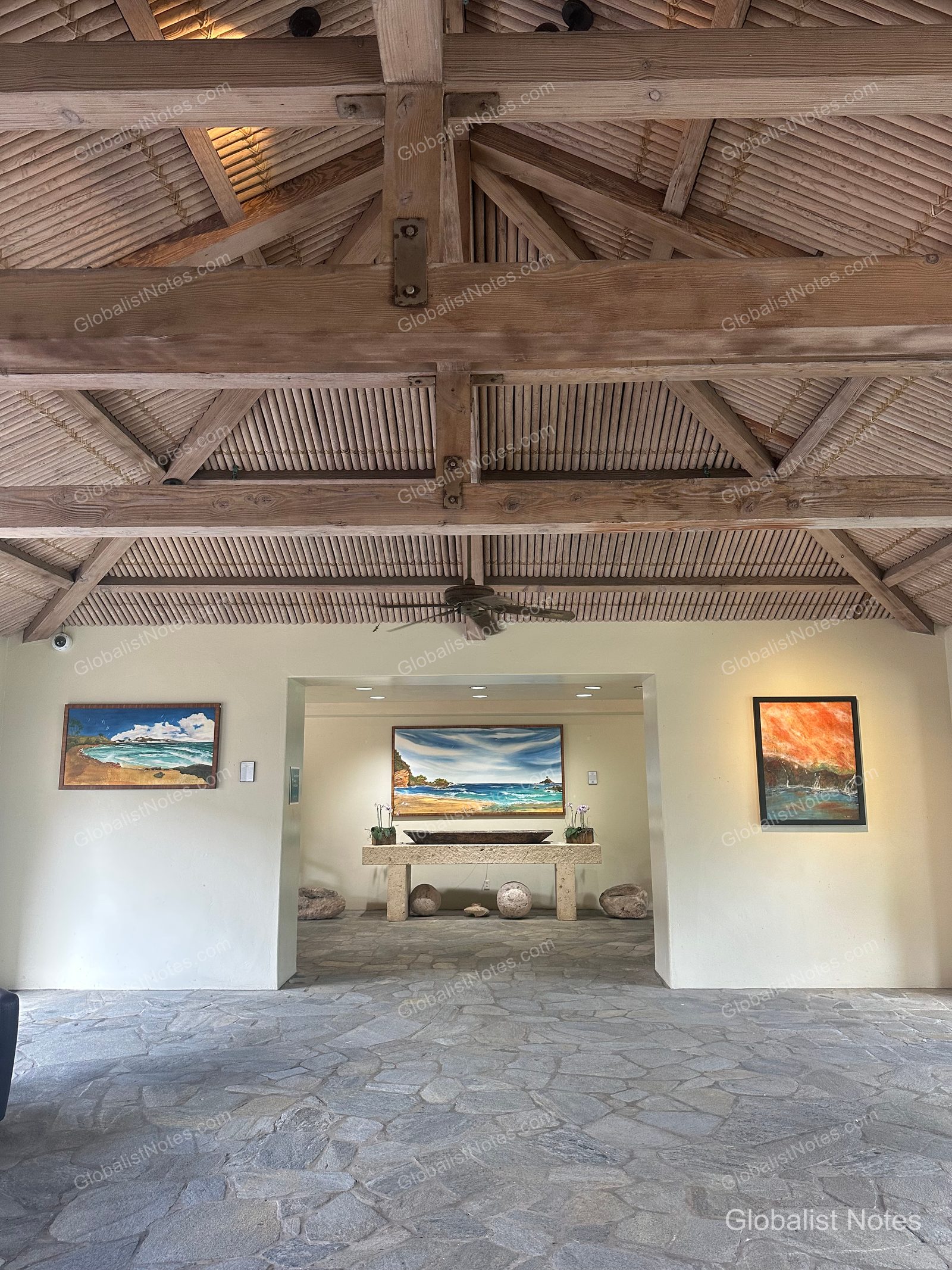 The lobby at Hana-Maui Resort features high wood-beamed ceilings and stone floors, with three oil paintings of Hawaiian beaches on the front and side walls and a long console table in the center.