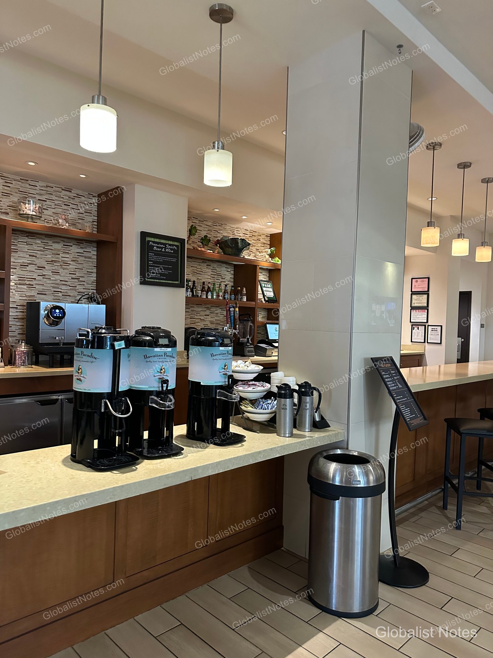Bar counter located on the lobby level of Hyatt Place Waikiki Beach. Three black coffee dispensers are lined up, with milk, sugar, and paper cups nearby. The back shelves display a liquor menu and bottles, and a counter table with bar stools is visible on the right.