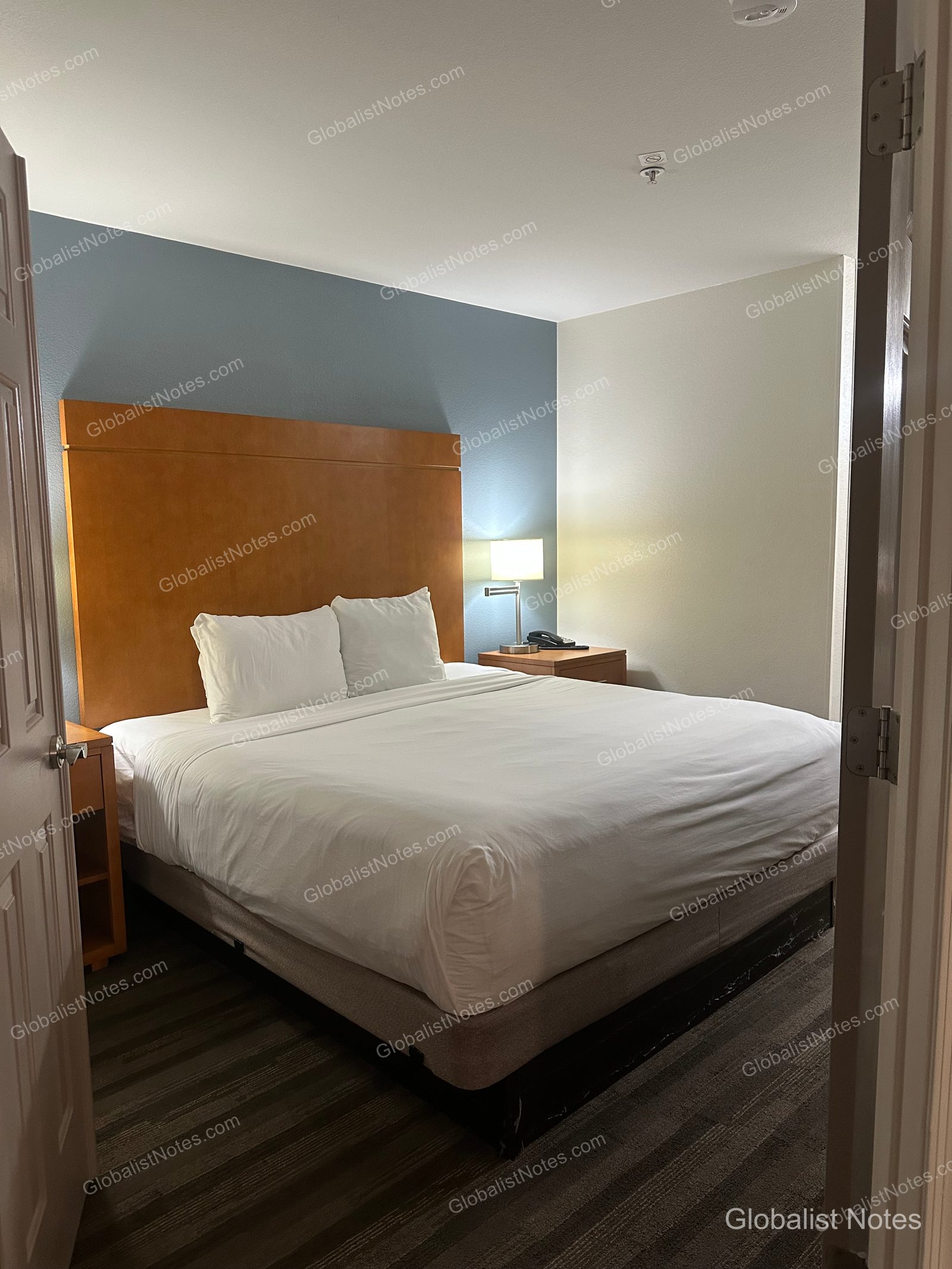 Hyatt House Scottsdale/Old Townの1ベッドルームスイートの寝室。ダークブルーのアクセントウォールを背景に、木製のヘッドボードを備えたキングサイズベッドが配置されています。