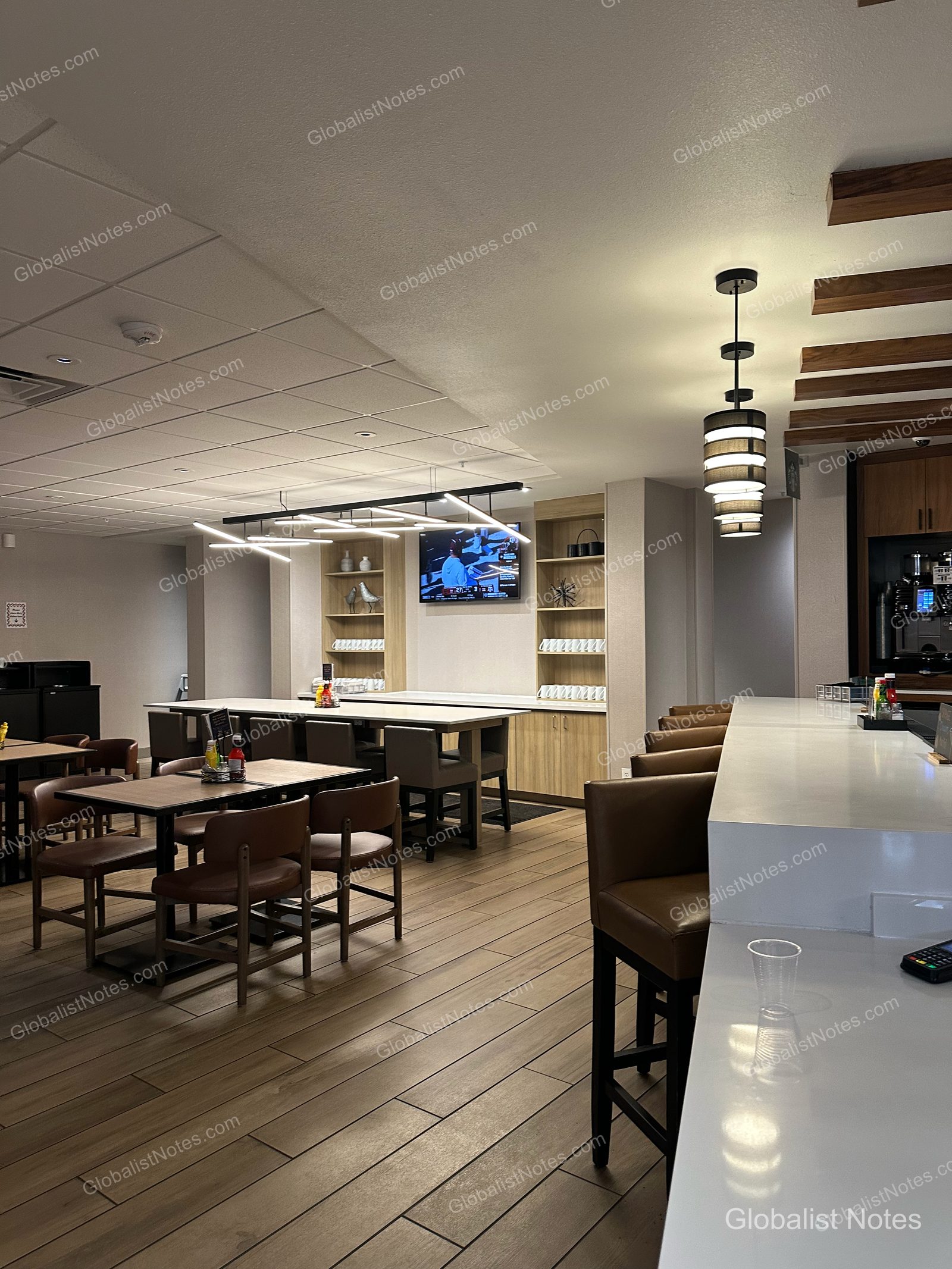 Hyatt Place Flagstaffの朝食エリア。木目調の床にテーブルと椅子が配置され、壁面には棚とテレビがある。右側にはハイチェア付きのカウンターがあり、天井にはモダンな照明が設置されている。