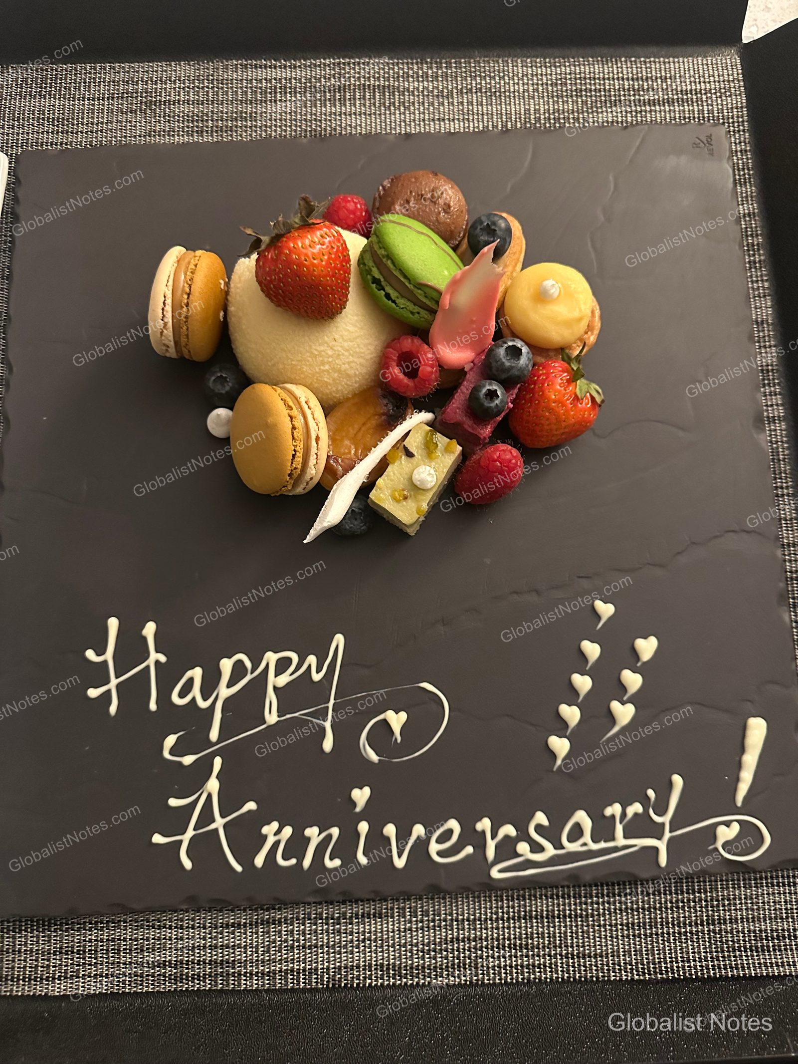 A black square plate holding fresh strawberries, raspberries, blueberries, and assorted colorful macarons, with 'Happy Anniversary!' and heart shapes written in white chocolate sauce at the bottom of the plate.