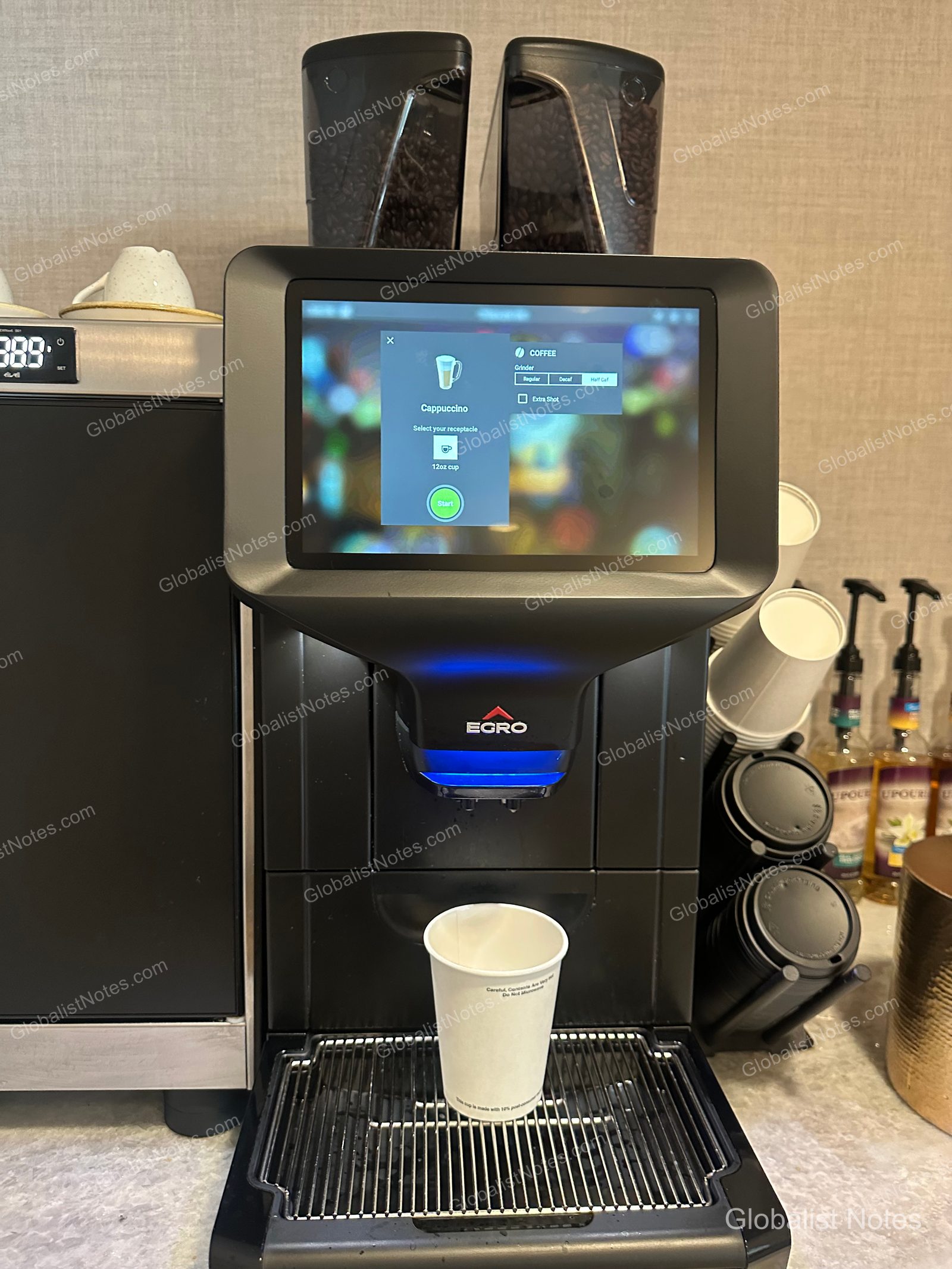 Egro automatic coffee machine in the Grand Hyatt San Antonio River Walk breakfast area. The touchscreen displays a cappuccino option and a 12oz cup setting, with disposable paper cups placed below.