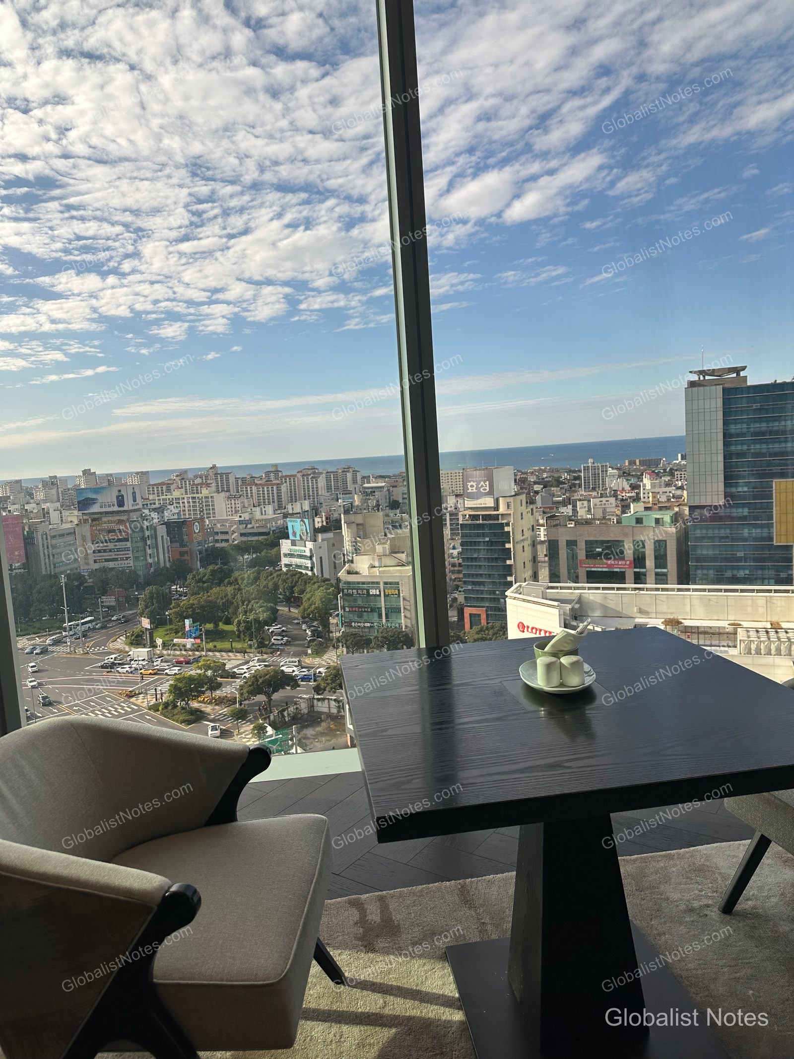 Floor-to-ceiling windows in the Grand Hyatt Jeju lounge overlook the Jeju city skyline and the distant blue ocean, with beige armchairs and black square tables arranged in the interior.