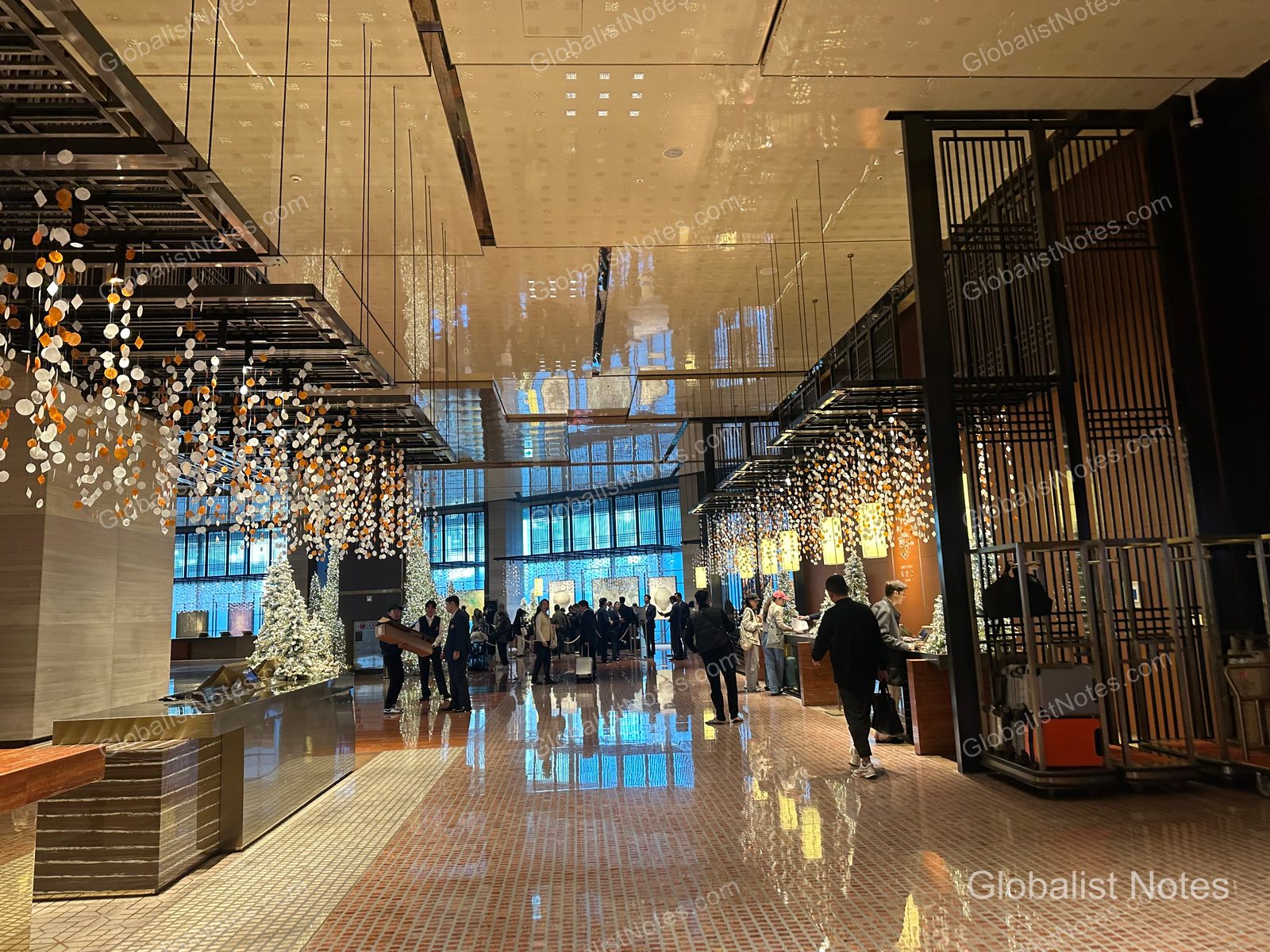 A view of the spacious lobby at Grand Hyatt Jeju, featuring numerous hanging light fixtures and Christmas tree decorations. The marble floor reflects the lighting, with guests waiting for check-in and hotel staff stationed throughout the lobby.