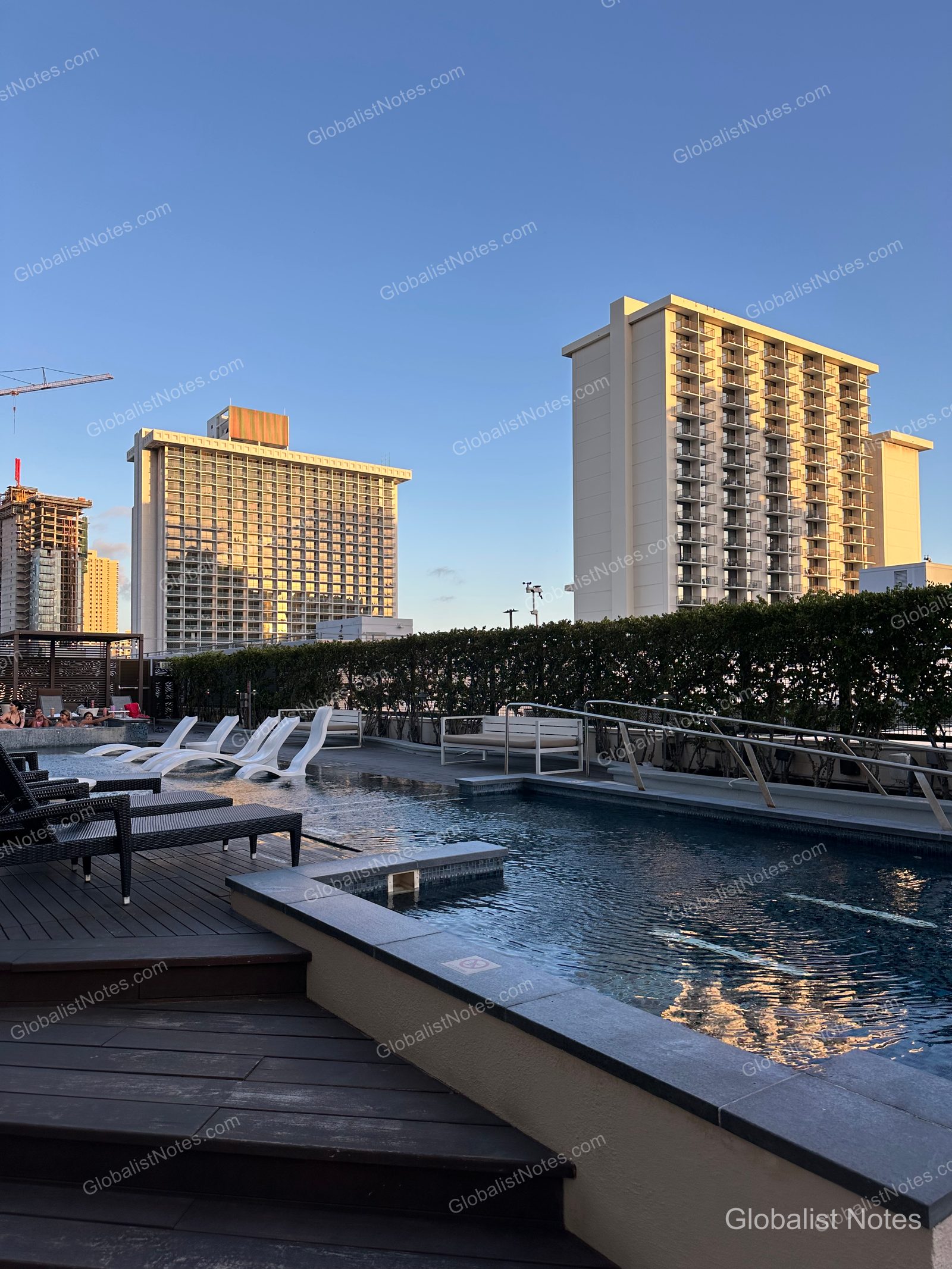 Outdoor pool deck at Hyatt Centric Waikiki Beach, featuring white sun loungers on a wooden deck with a view of the sunset-lit Honolulu skyline in the background.
