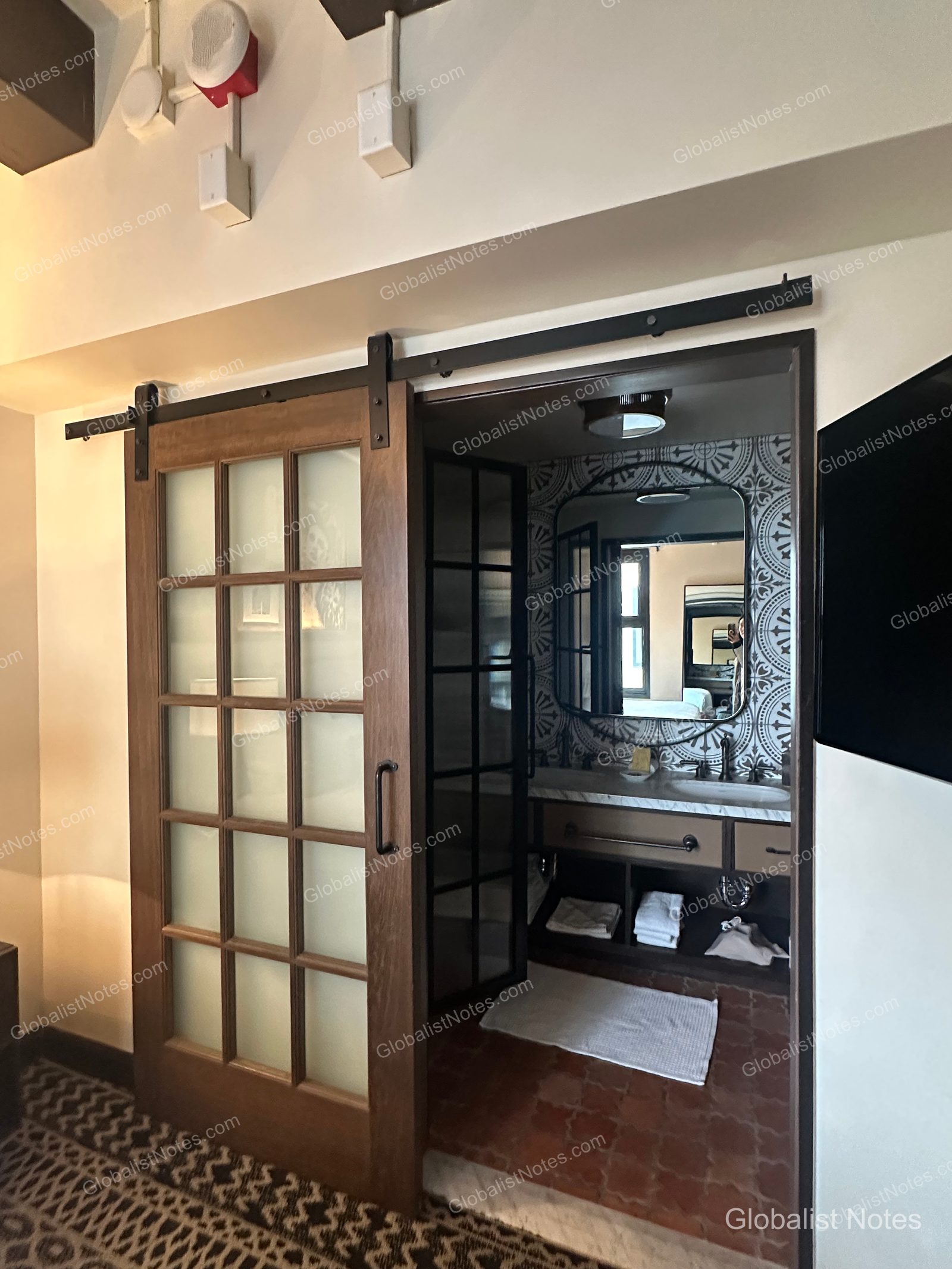 Bathroom entrance of the Junior Suite at Hotel Figueroa, Los Angeles. A wooden-framed lattice sliding door is partially open, revealing geometric patterned tile walls, a vanity with a mirror, and neatly stacked towels in the lower storage area.
