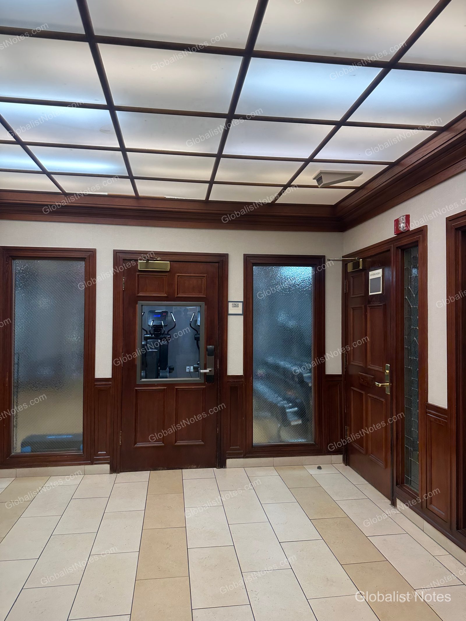 Hyatt House Scottsdale/Old Townのフィットネスセンター入り口。格子状の天井照明の下にダークウッドフレームのガラスドアが並び、奥にトレーニングマシンが一部見える。