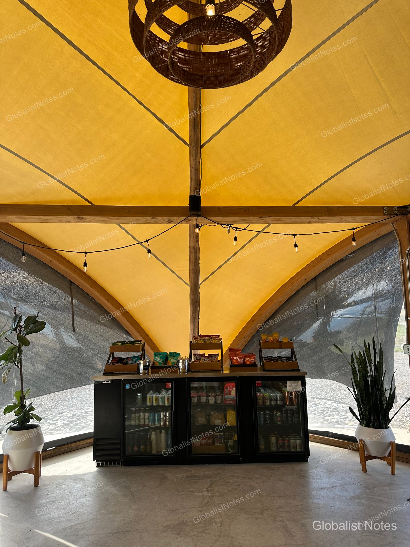 Under Canvas North Yellowstoneのテント内部。中央の棚にはドリンクやスナックが並び、黄色いキャンバス地の天井には照明が吊るされています。両サイドには観葉植物が置かれています。