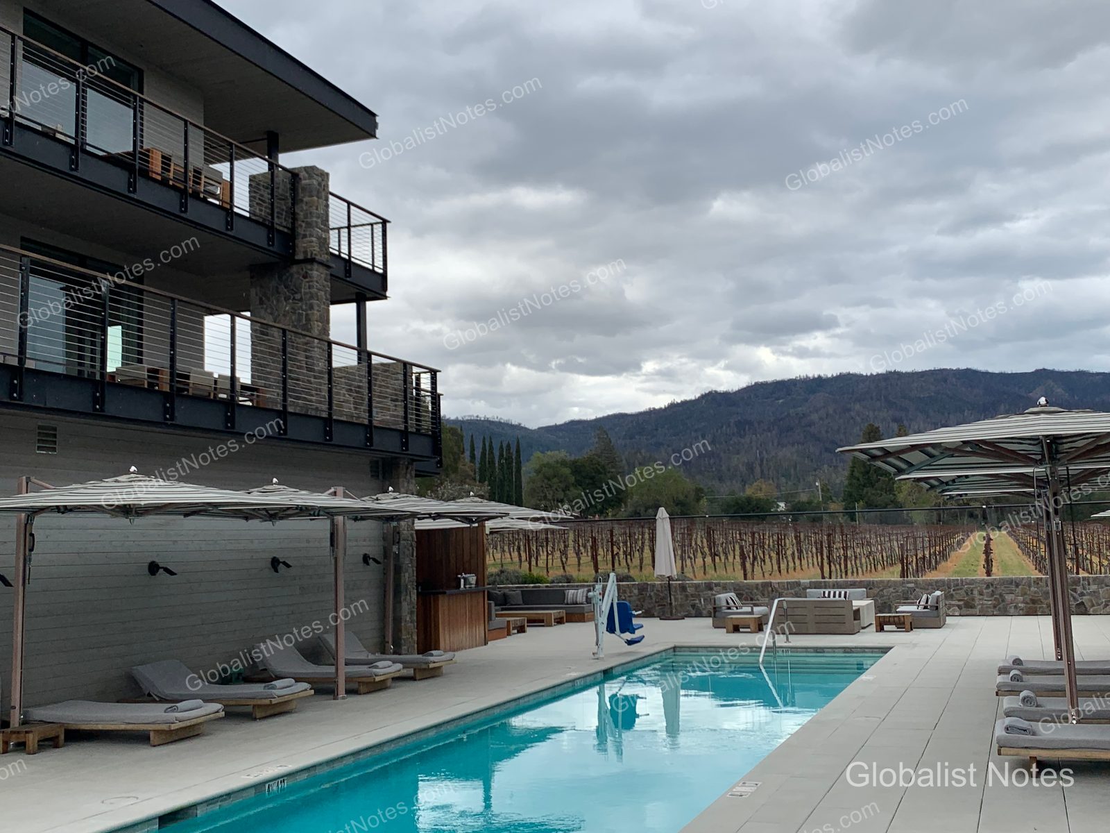 Outdoor pool at Alila Napa Valley in St. Helena, California. A rectangular pool sits beside the modern hotel building, with umbrellas and sun loungers arranged on the deck. In the background, Napa Valley vineyards and low mountain ranges are visible under a cloudy sky.