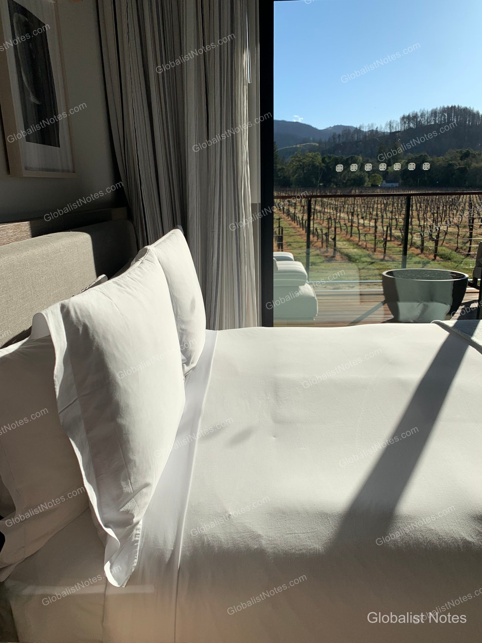View from inside an Alila Napa Valley guestroom, showing a queen bed with white linens next to floor-to-ceiling windows that open to a balcony overlooking vineyards and mountains.