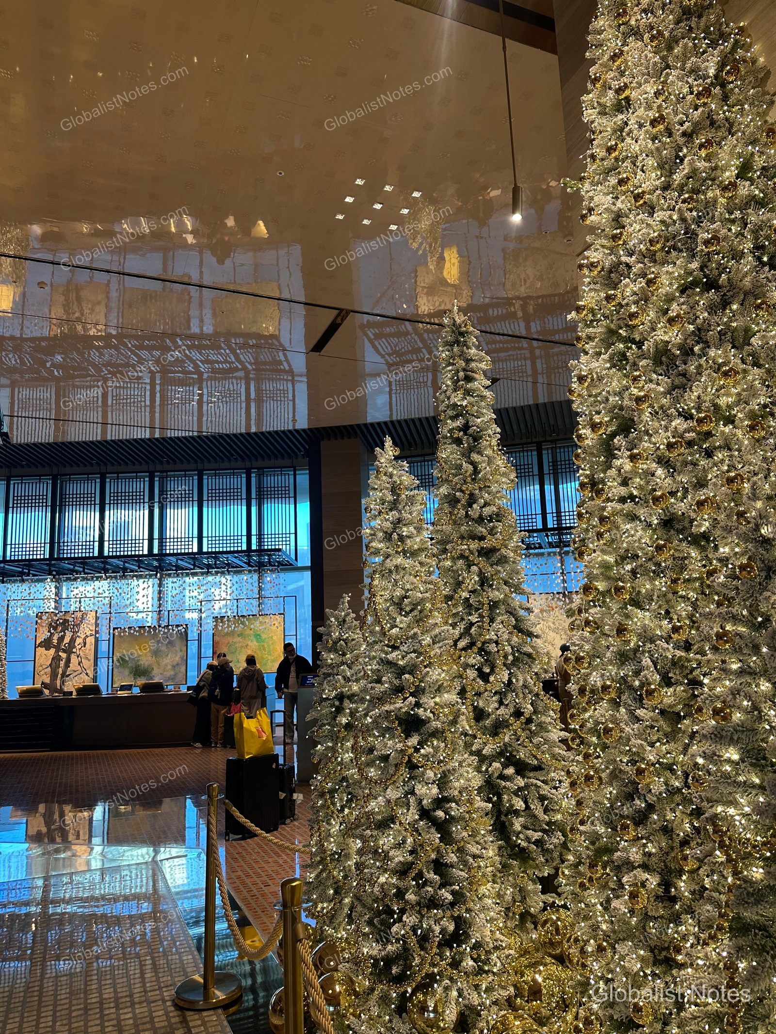 Multiple tall Christmas trees decorated with gold ornaments stand in the Grand Hyatt Jeju lobby, with the check-in counters and modern art installations visible in the spacious area behind them.